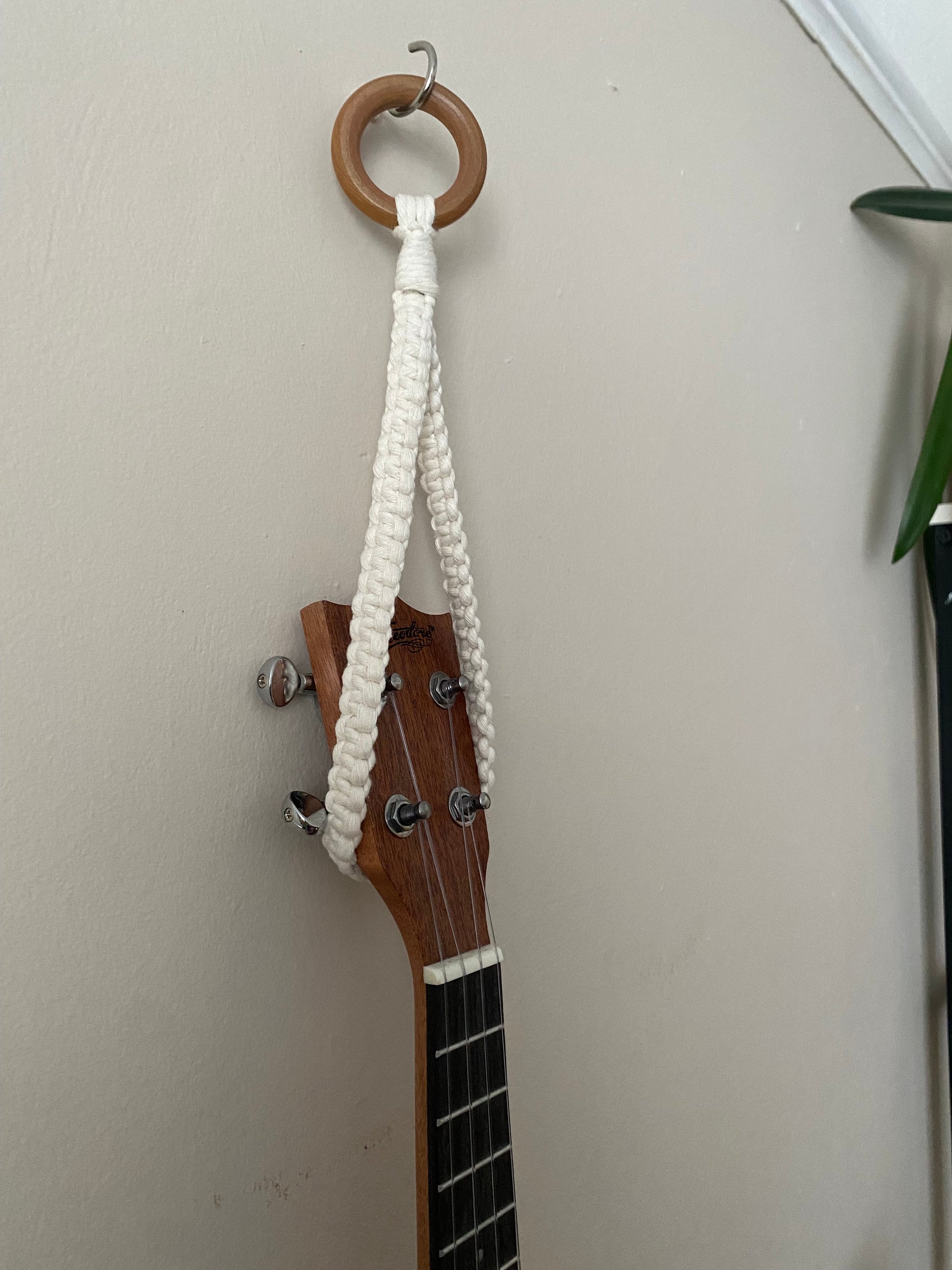 Close up of the headstock of a dark brown ukulele, hanging against a beige wall from a simple macrame knotted loop hanger, made with cotton yarn and a wooden ring.