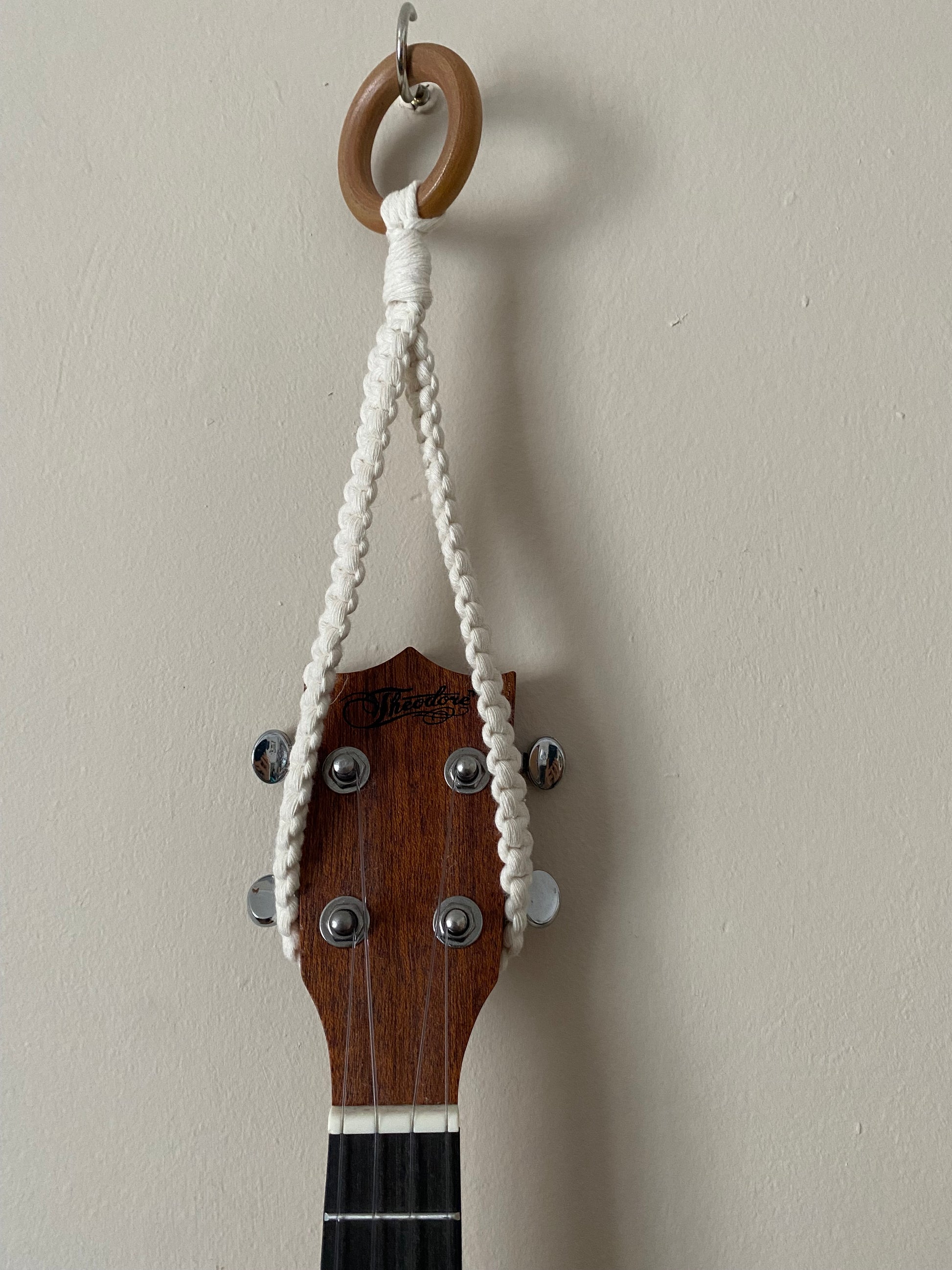Close up of the headstock of a dark brown ukulele, hanging against a beige wall from a simple macrame knotted loop hanger, made with cotton yarn and a wooden ring.