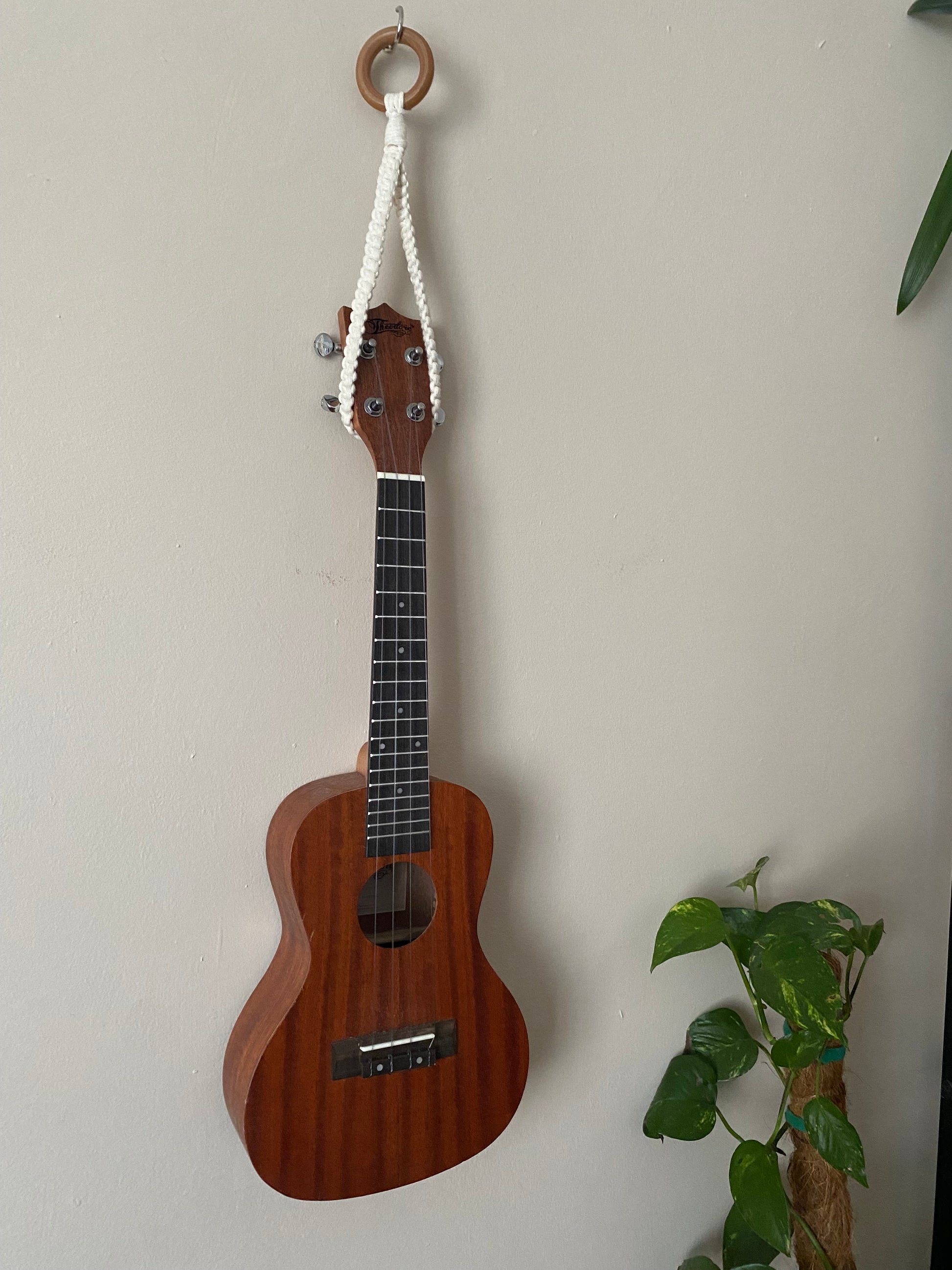 A dark brown ukulele, hanging against a beige wall from a simple macrame knotted loop hanger, hooked around the headstock of the instrument, made with cotton yarn and a wooden ring.