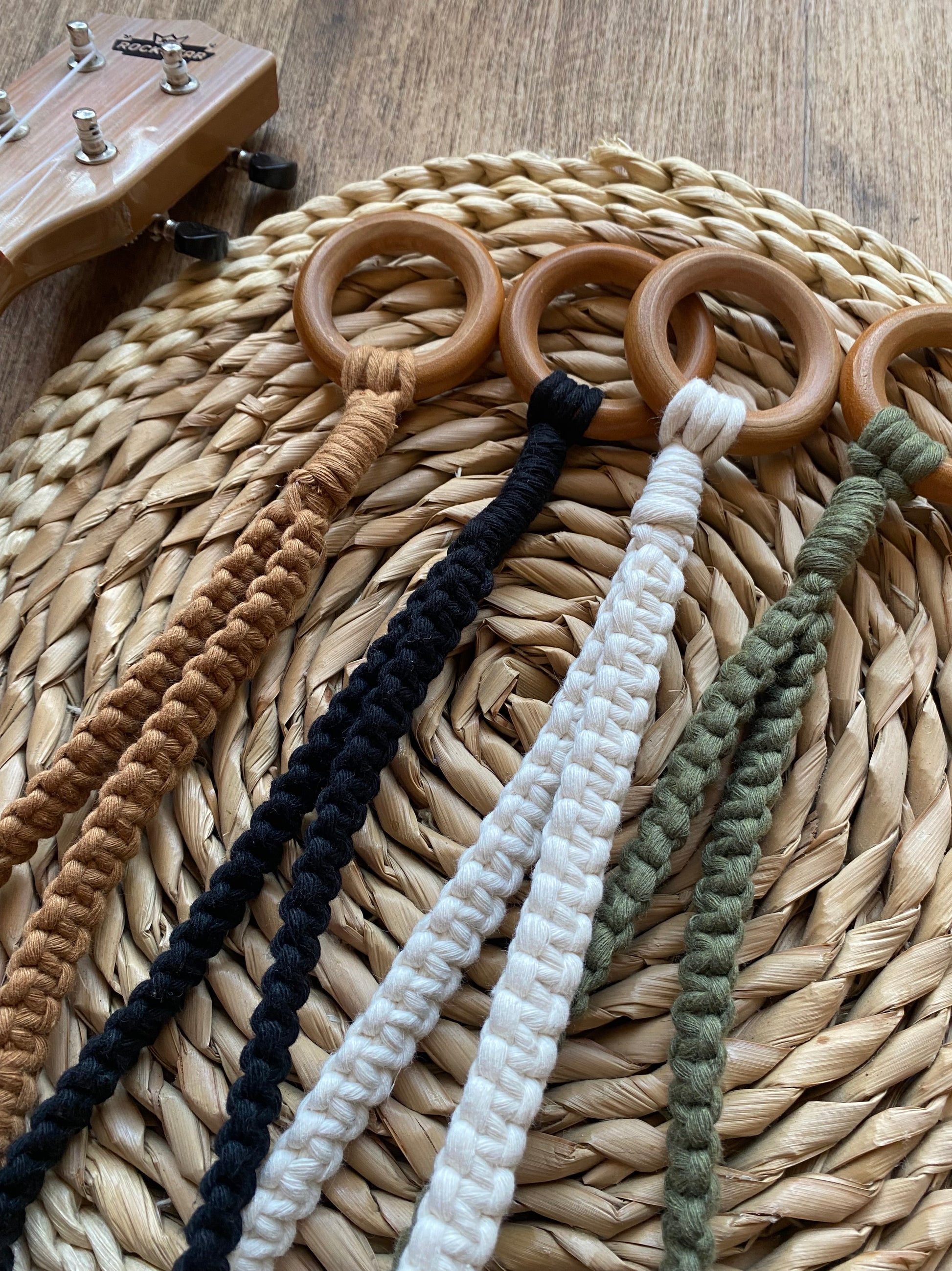 Four handmade macrame loop ukulele hangers made with brown, black, natural and green yarns, each topped with a wooden ring, laid next to each other on a wicker mat. The head of a light brown ukulele is just sign to the left of the image.