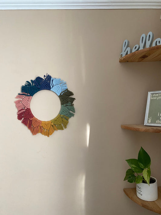 A macrame mandala wall hanging made in rainbow colours, hangs on a beige wall next to a set of wooden corner shelves.