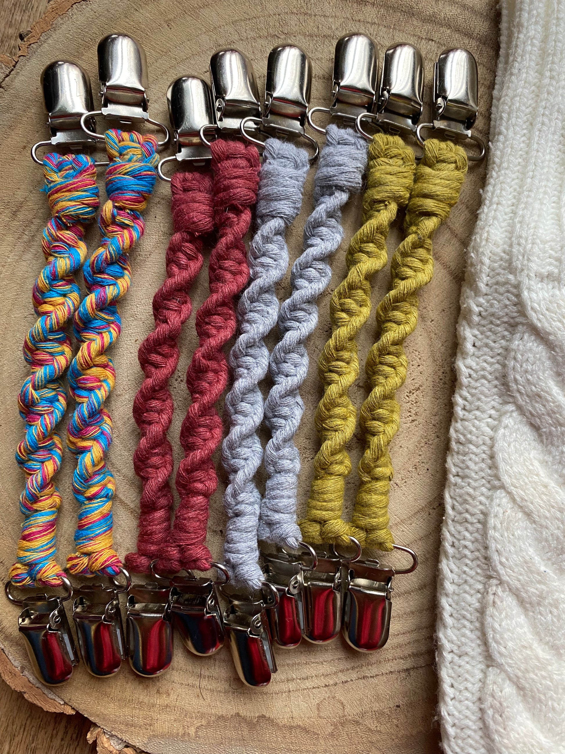 Four pairs of handmade macrame mitten clips made with a twisted knot pattern, with metal clips on each end, laid out on a wooden board.