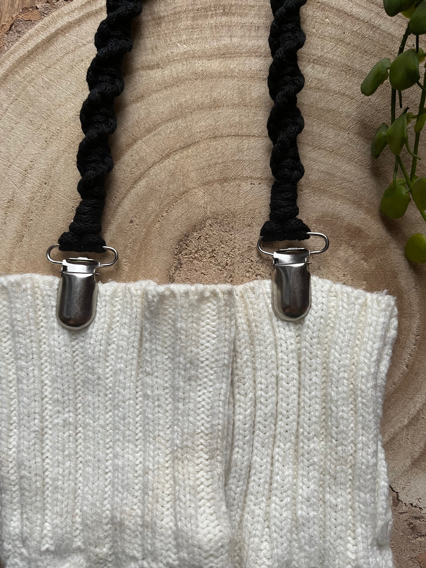 A pair of handmade macrame mitten clips made with black yarn with metal clips on the end, attached to a pair of white mittens on a wooden board.