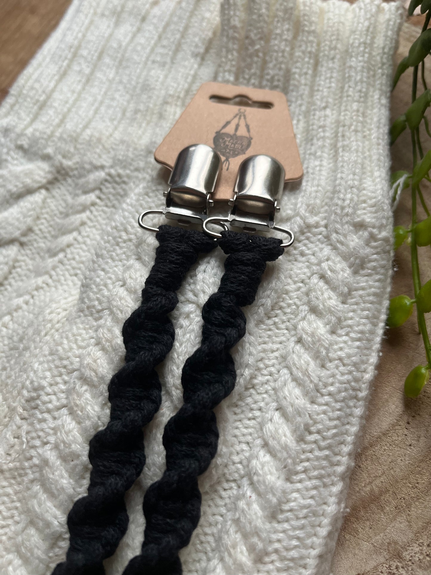 A pair of handmade macrame mitten clips made with black yarn with metal clips on the end, attached to a cardboard tag and displayed on top of a pair of white mittens on a wooden board.