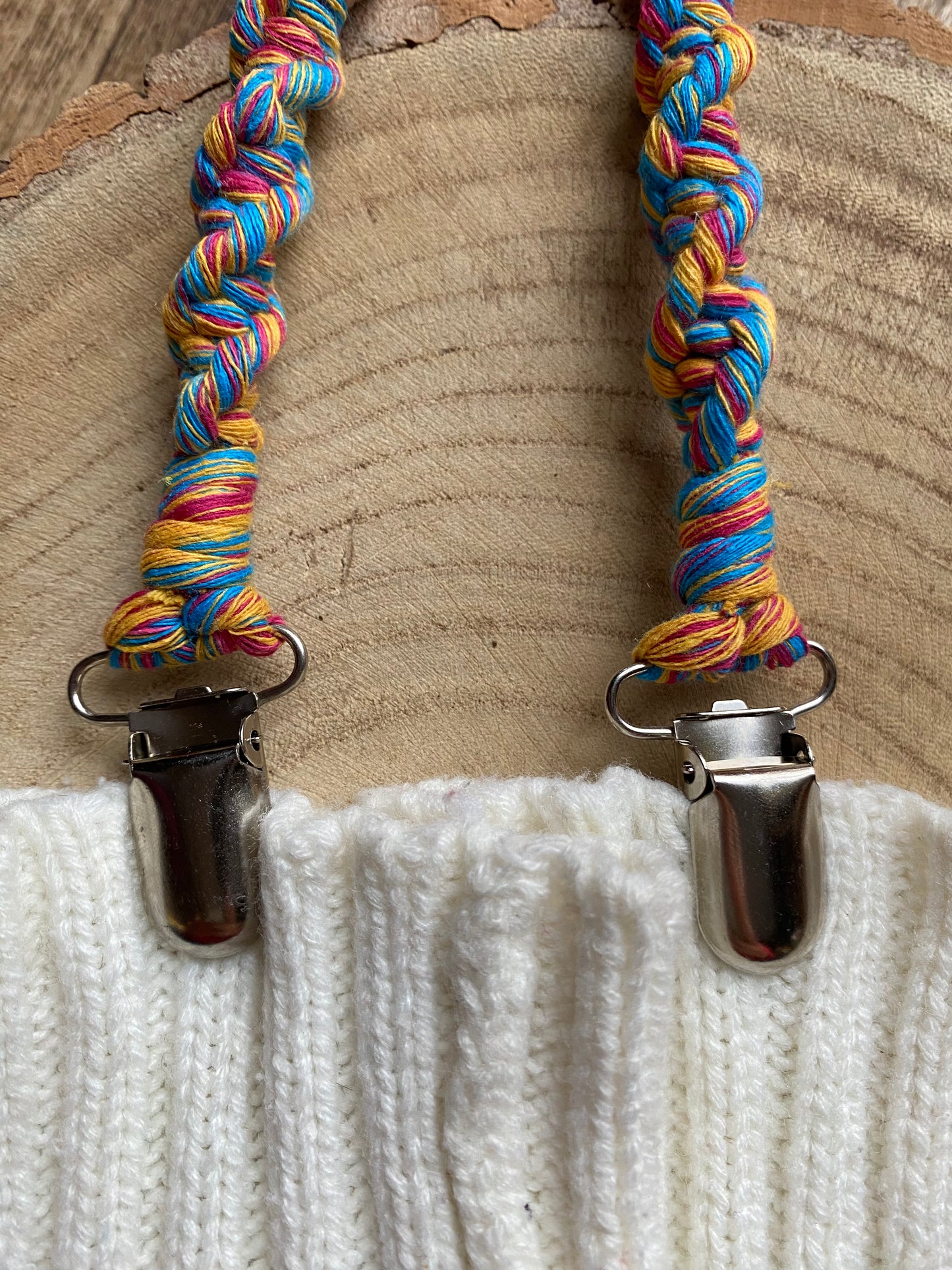 A pair of handmade macrame mitten clips made with multicoloured yarn with metal clips on the end, attached to a pair of white mittens on a wooden board.