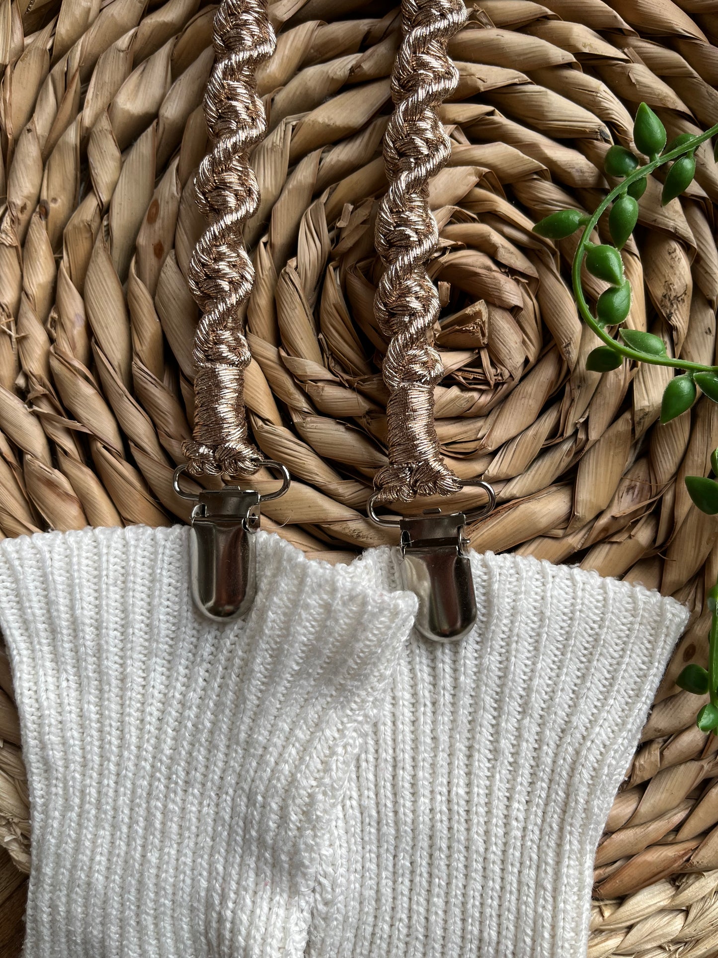 A pair of handmade macrame mitten clips made with metallic yarn with metal clips on the end, attached to a pair of white mittens on a wicker board.