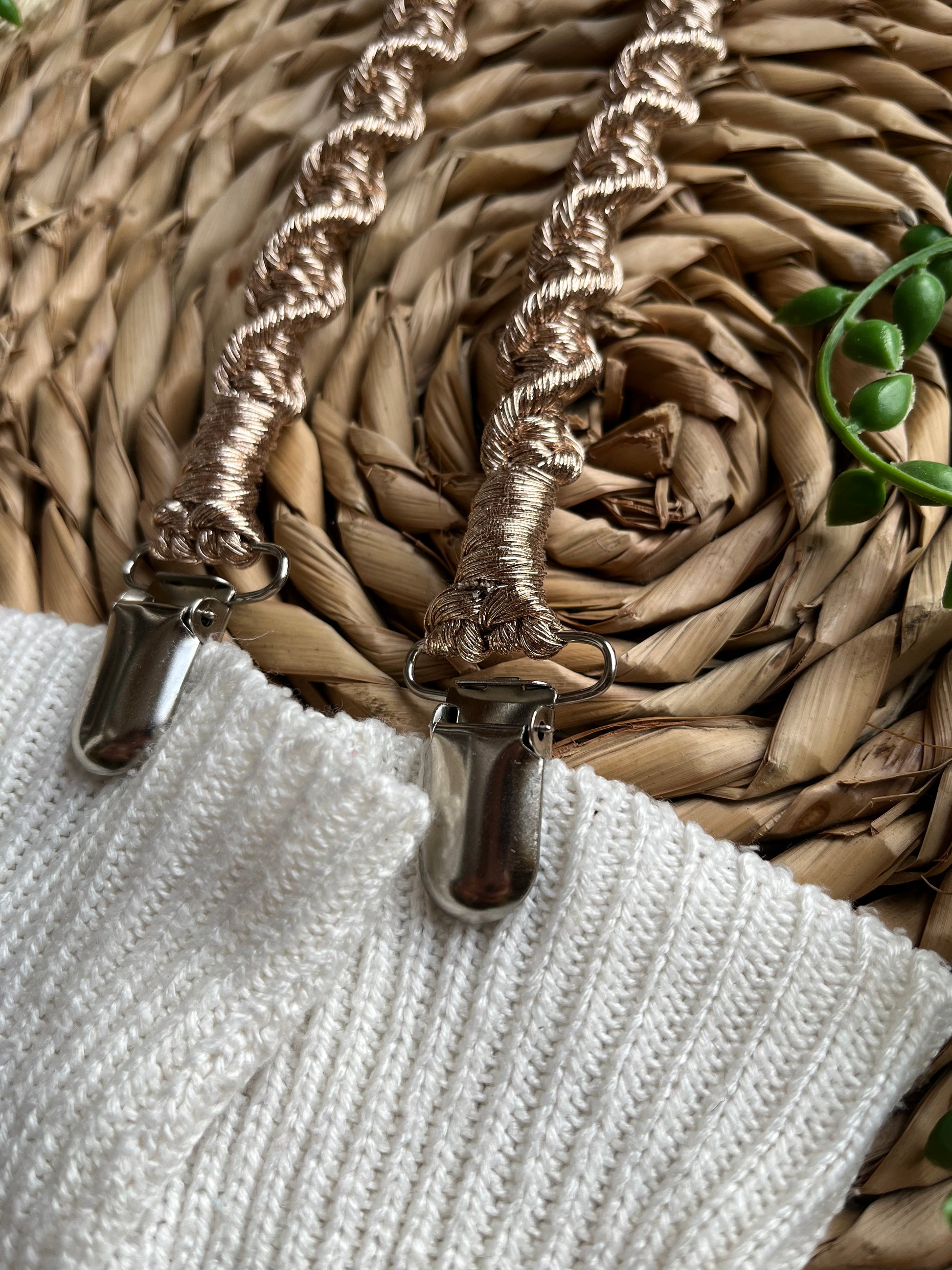 Close up of a pair of handmade macrame mitten clips made with metallic gold yarn with metal clips on the end, attached to a pair of white mittens on a wicker mat.
