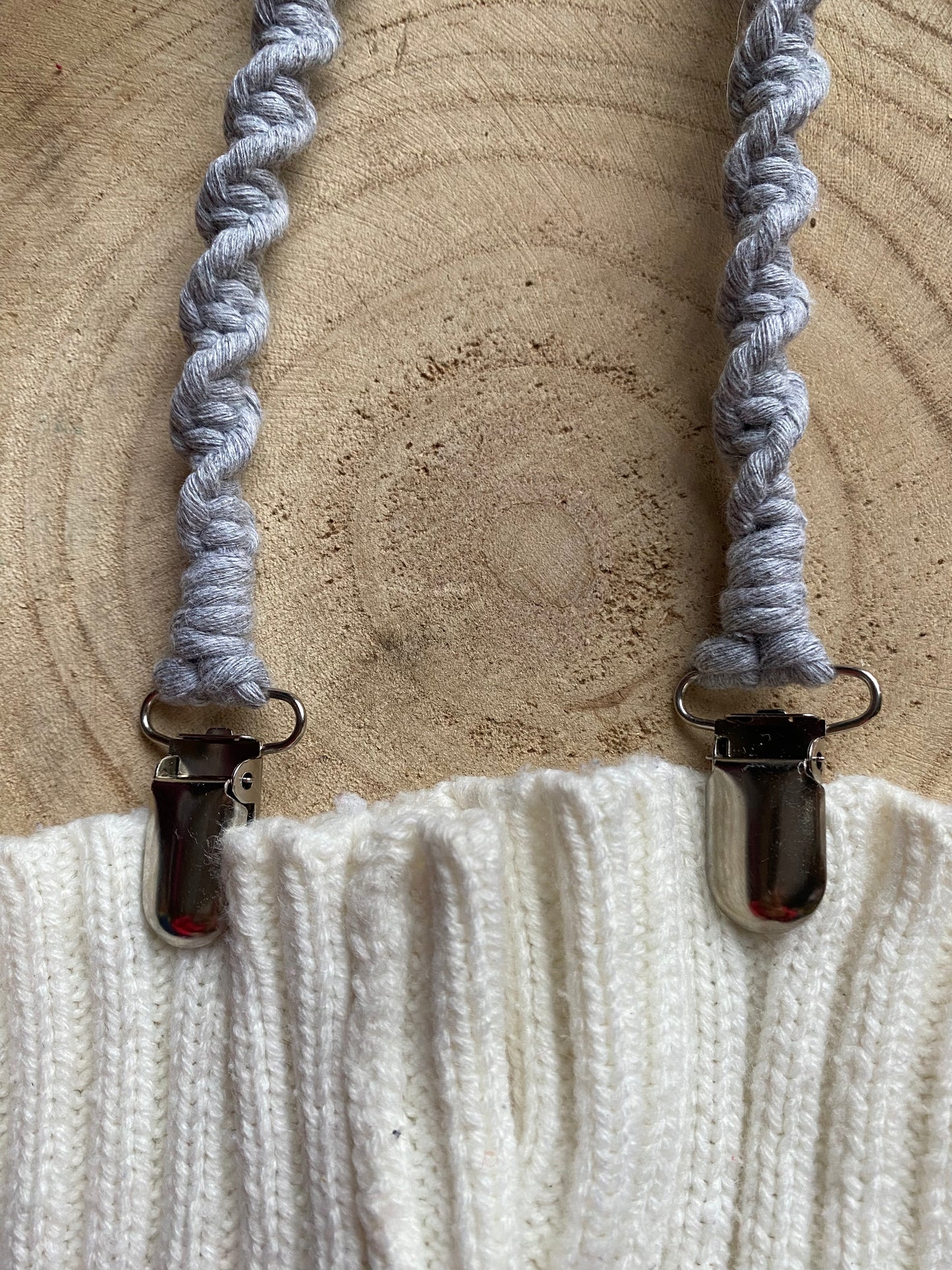 A pair of handmade macrame mitten clips made with grey yarn with metal clips on the end, attached to a pair of white mittens on a wooden board.