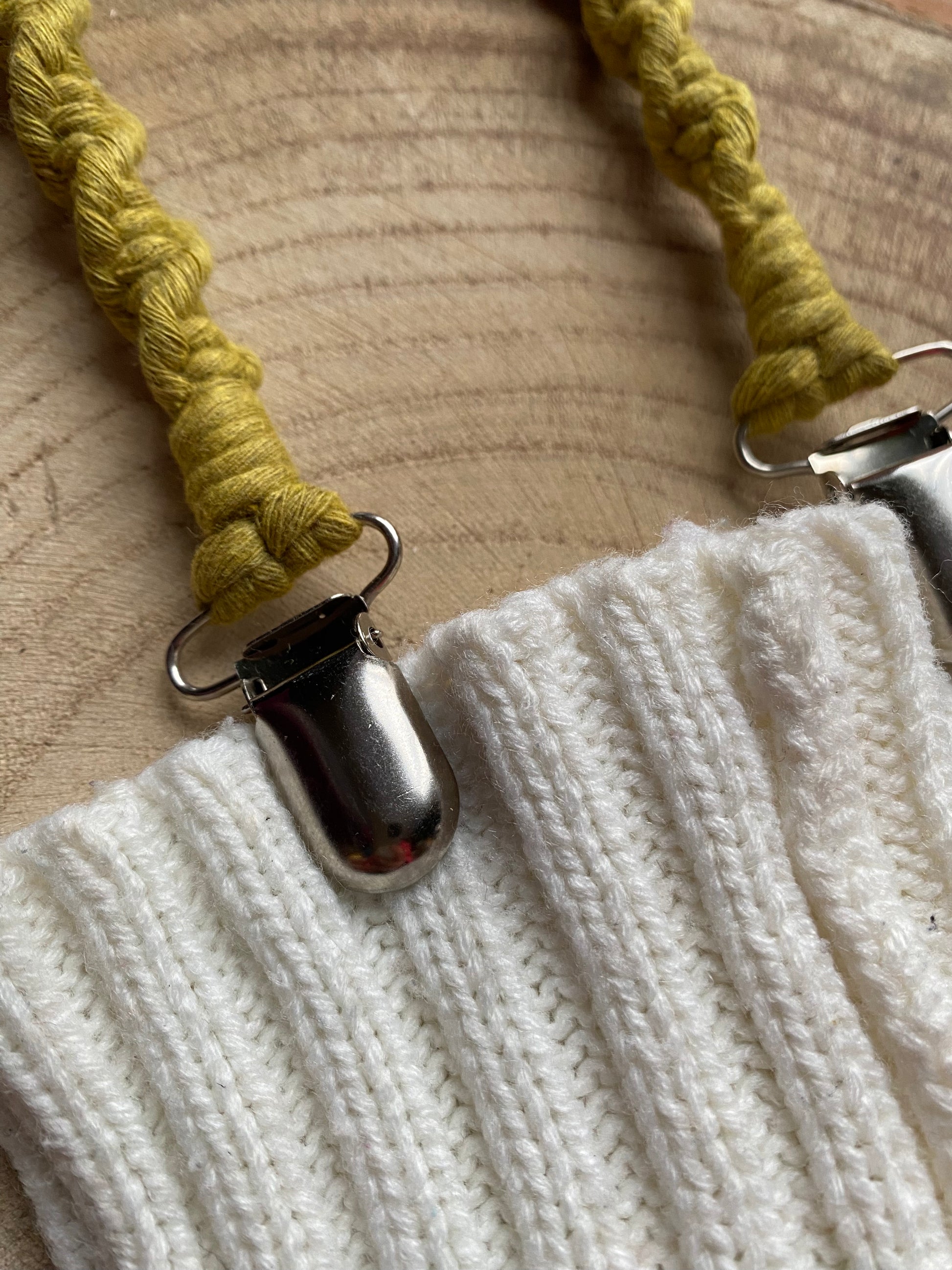 A pair of handmade macrame mitten clips made with bright green yarn with metal clips on the end, attached to a pair of white mittens on a wooden board.
