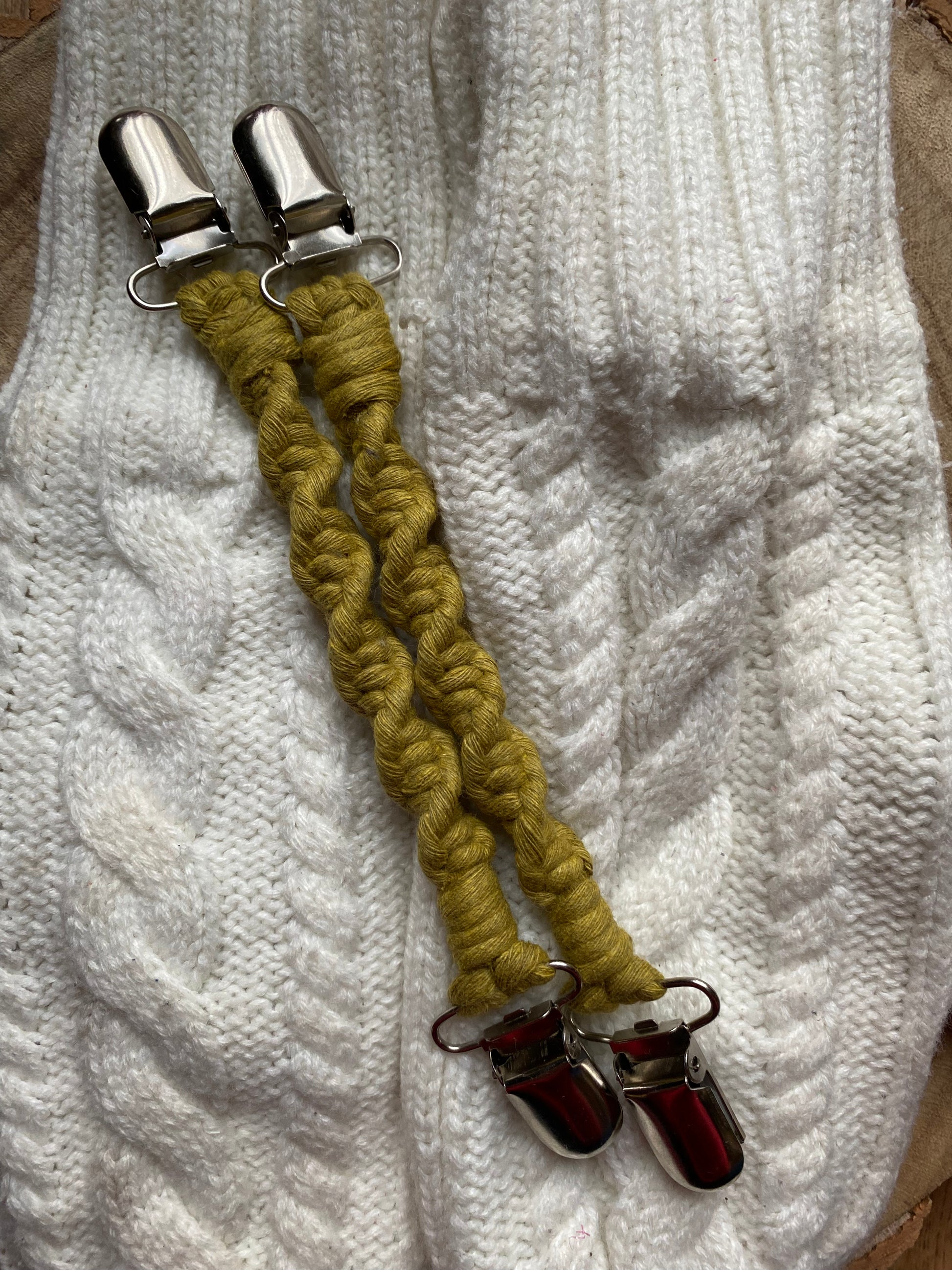 A pair of handmade macrame mitten clips made with bright green yarn with metal clips on each end, laying on top of a pair of white mittens.