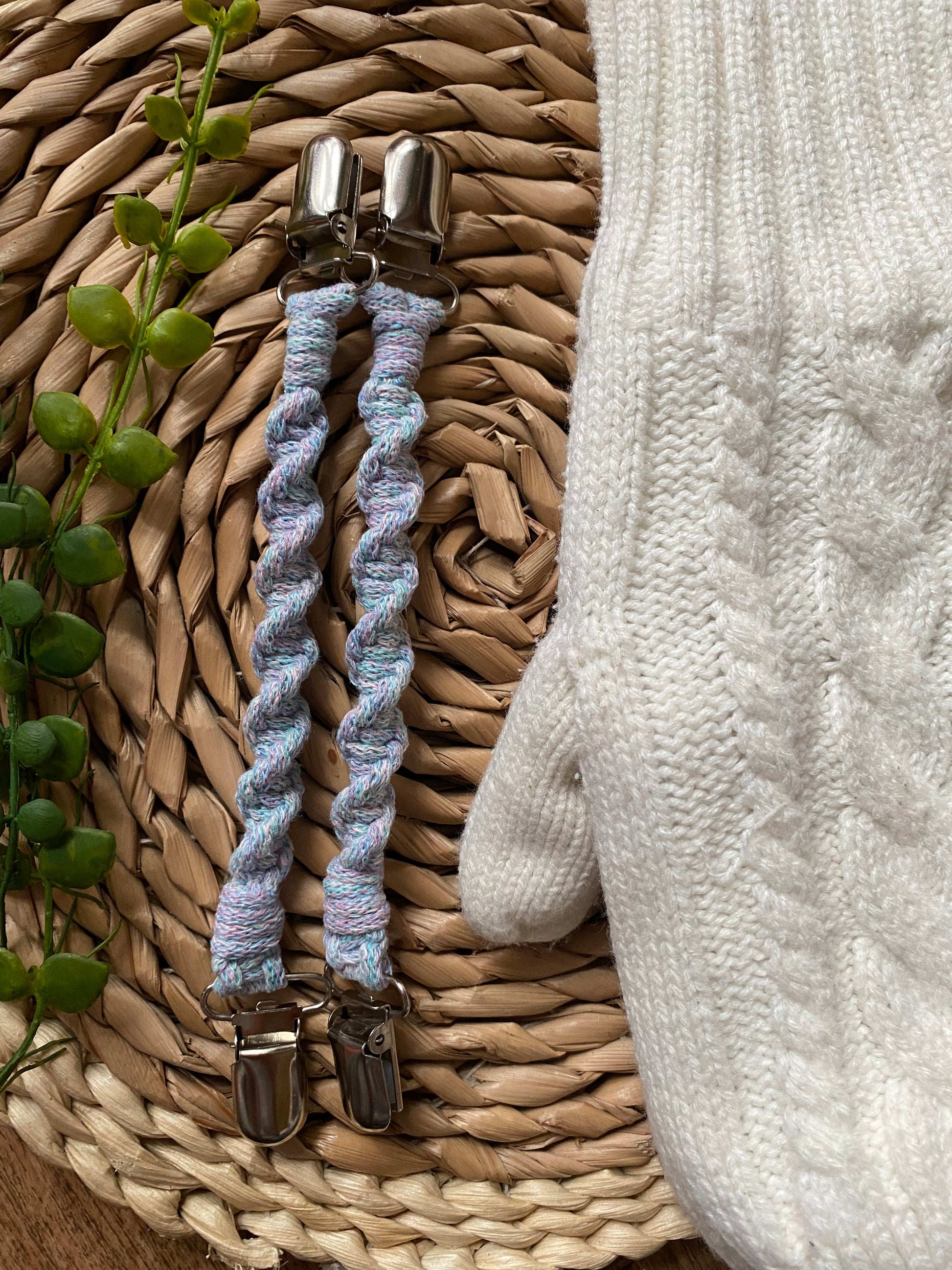 A pair of handmade macrame mitten clips made with pastel yarn with metal clips on each end, laid on wicker mat next to a pair of white mittens.