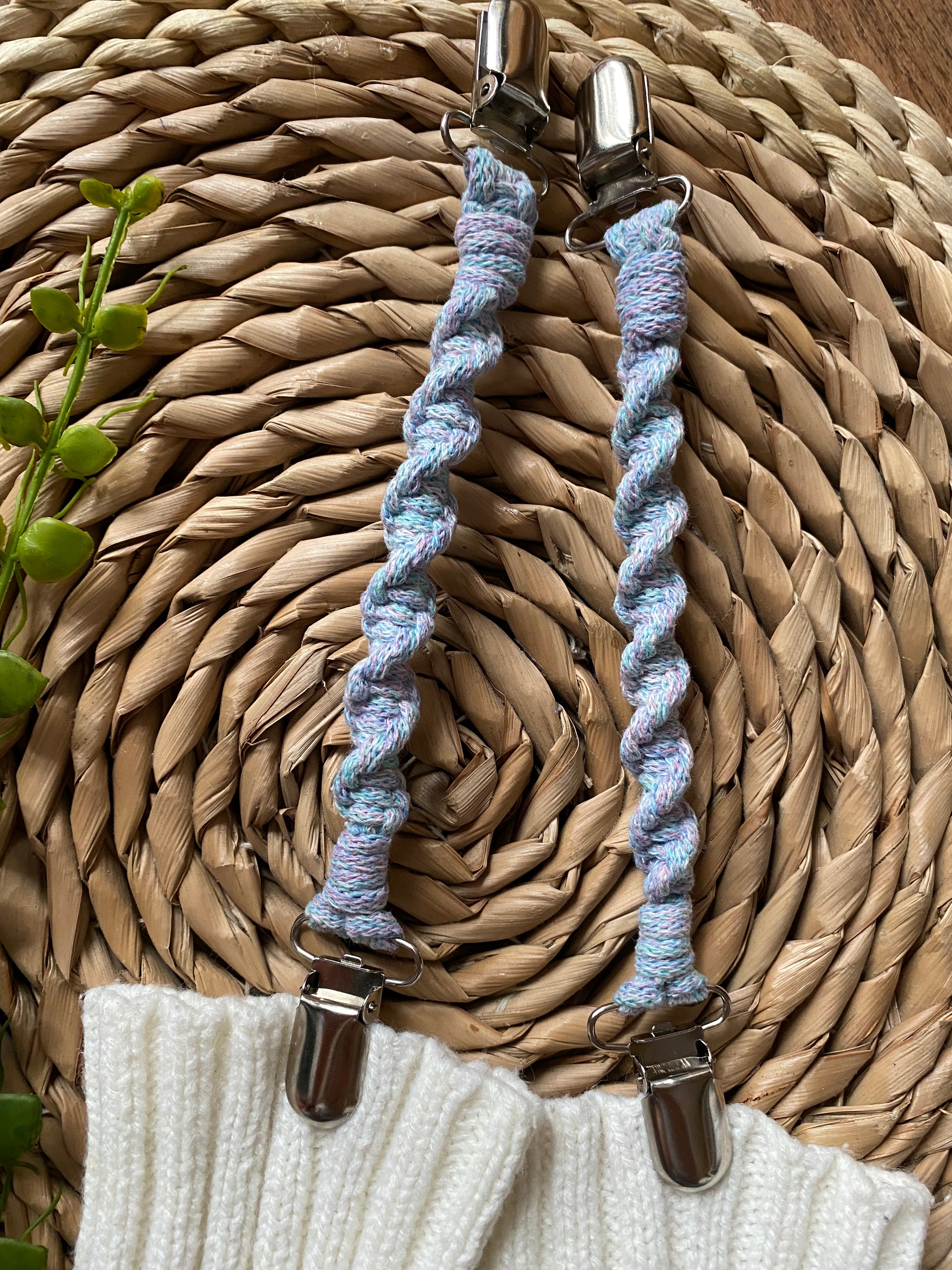 A pair of handmade macrame mitten clips made with pastel yarn with metal clips on each end, attached to a pair of white mittens on a wicker board.