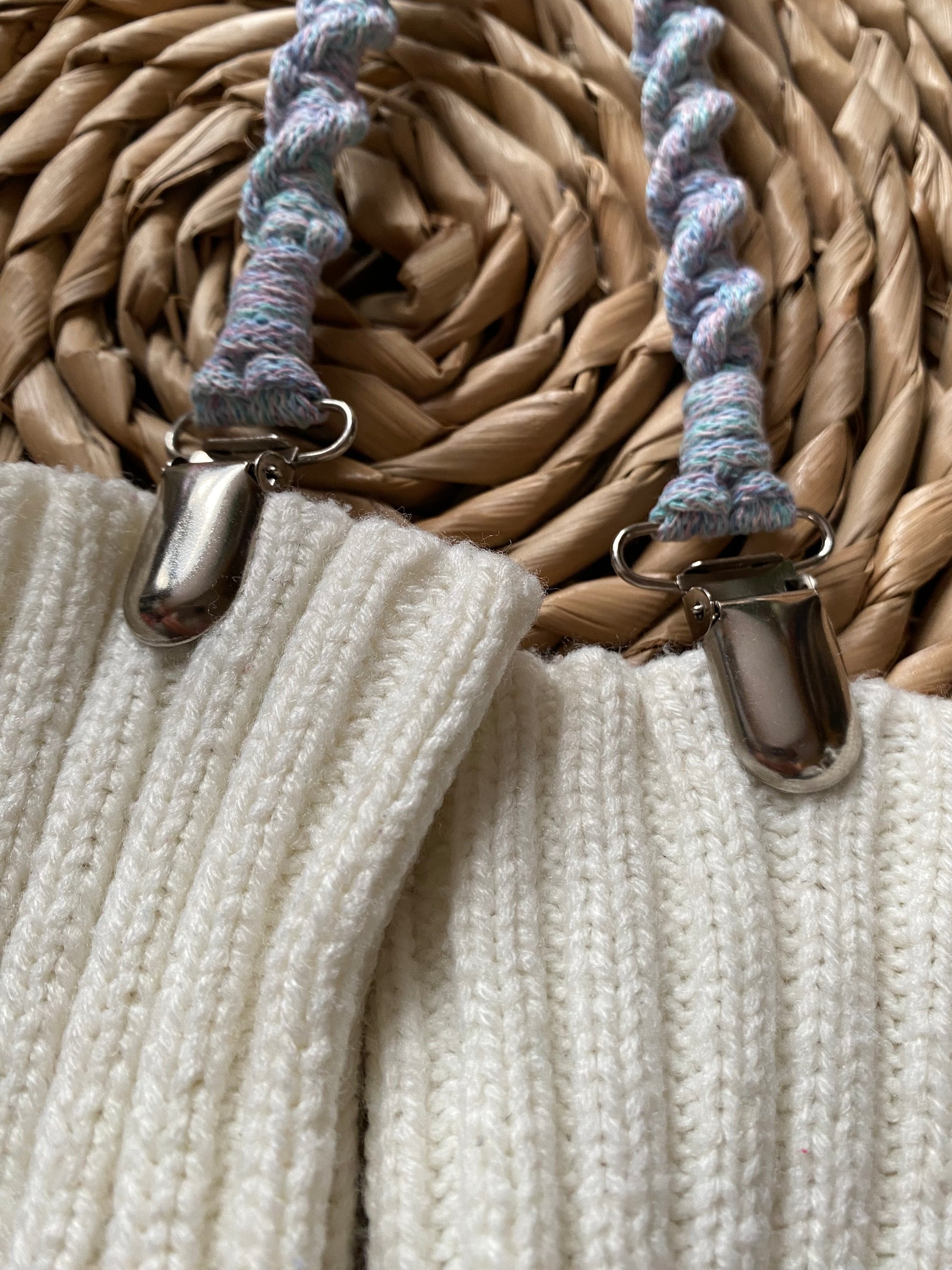 Close up of a pair of handmade macrame mitten clips made with pastel yarn with metal clips on the end, attached to a pair of white mittens on a wicker board.
