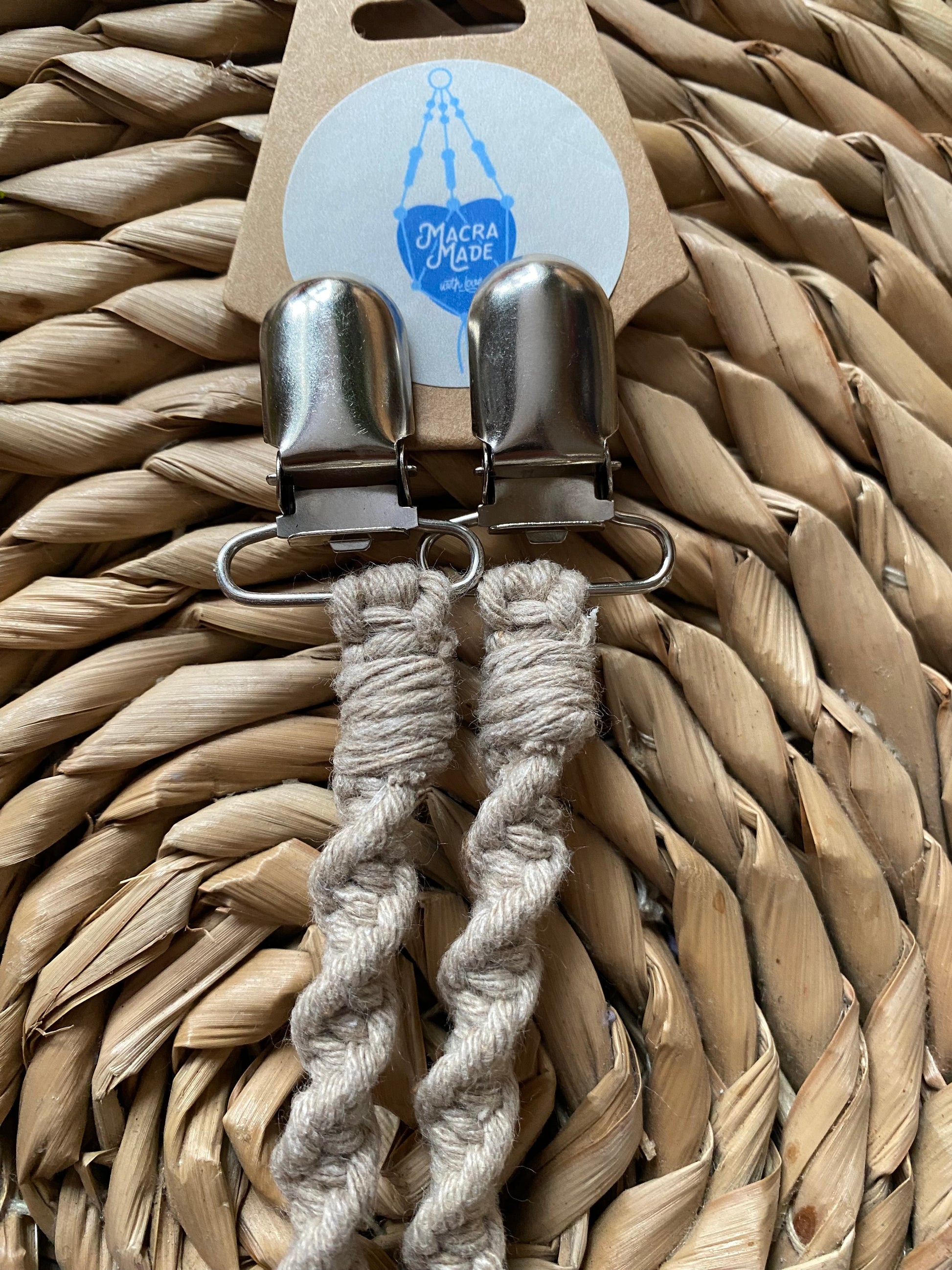 A pair of handmade macrame mitten clips made with taupe yarn with metal clips on the end, attached to a cardboard tag, on a wicker mat.