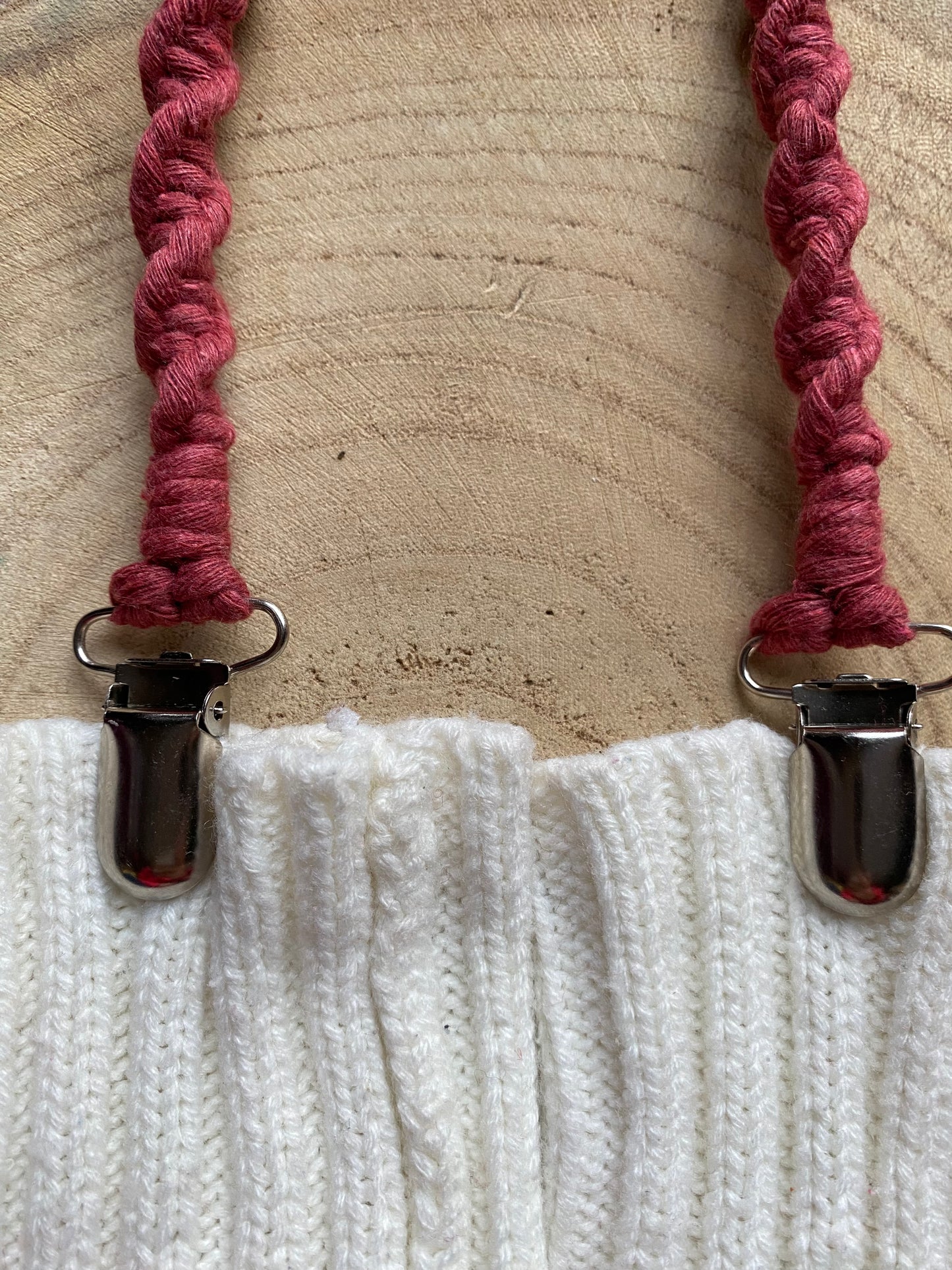A pair of handmade macrame mitten clips made with dark pink yarn with metal clips on the end, attached to a pair of white mittens on a wooden board.