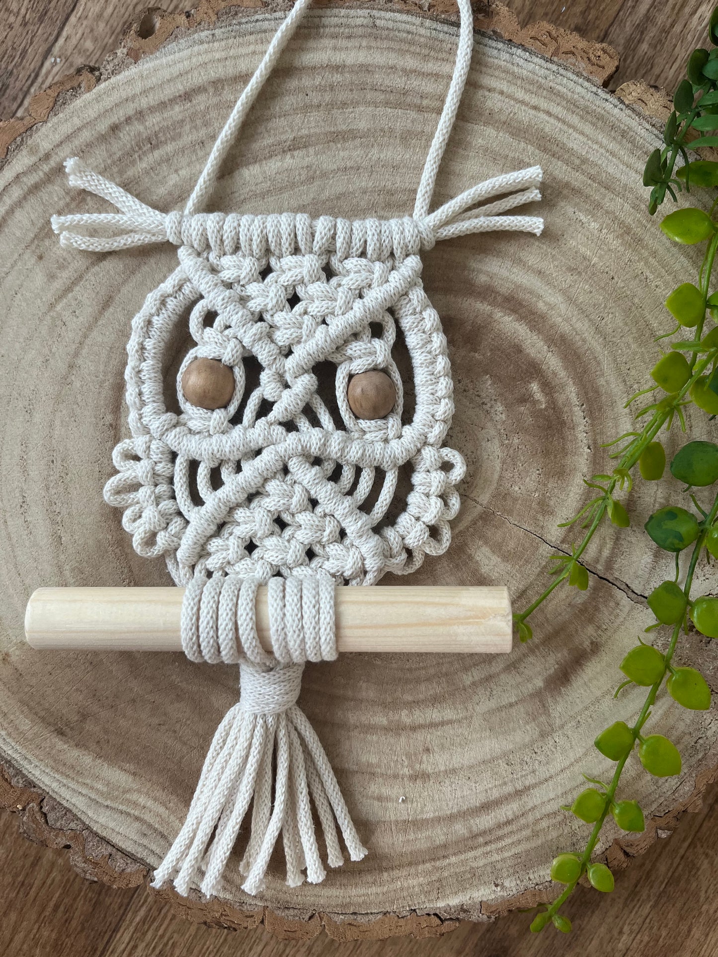 Handmade macrame owl wall hanging decoration, perched on a wooden dowel and displayed flat on a wooden board. Made with natural cotton and wooden beads.