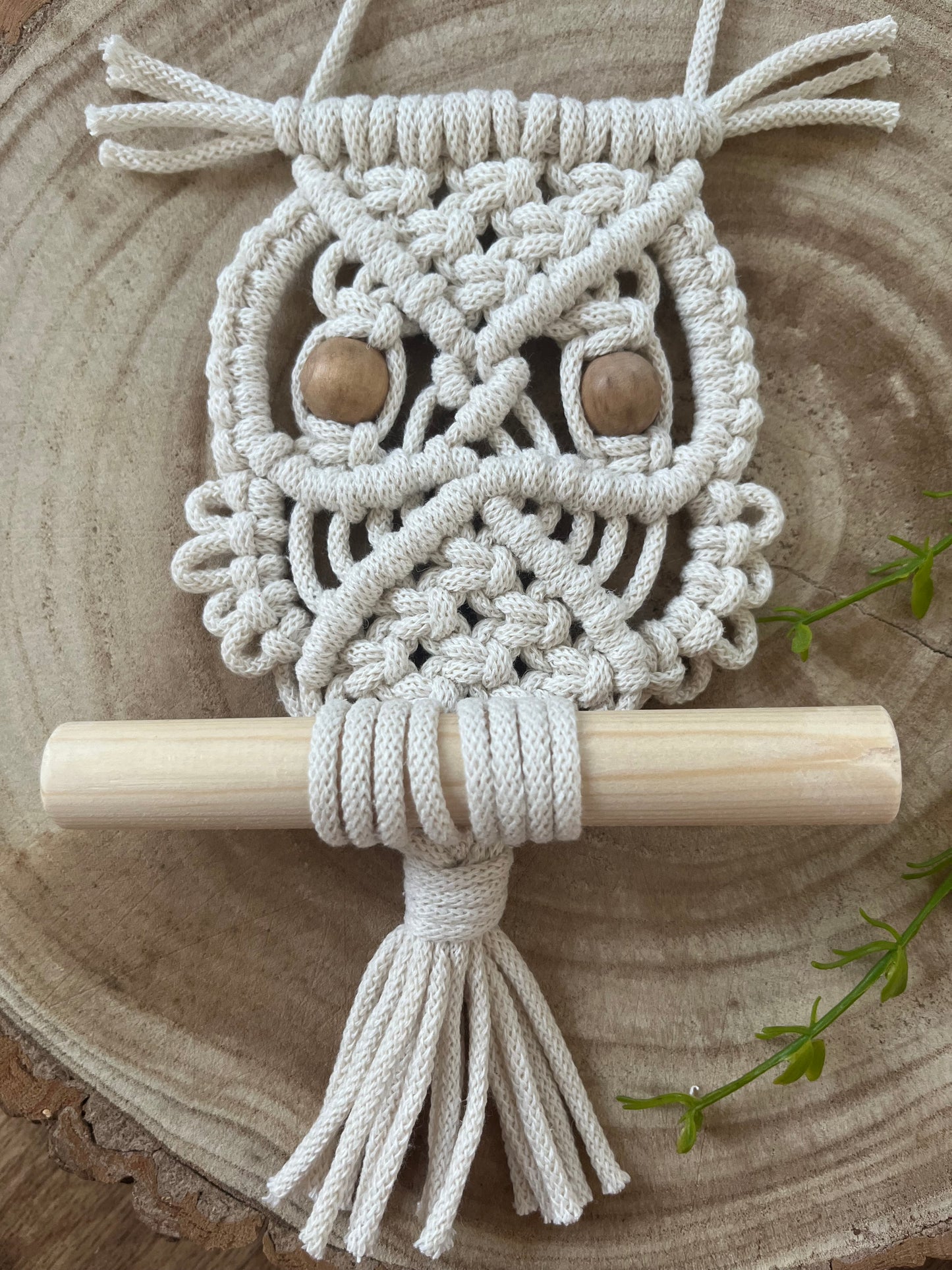 Close up of a handmade macrame owl wall hanging decoration, perched on a wooden dowel. Made with natural cotton and wooden beads, displayed on a wooden board.