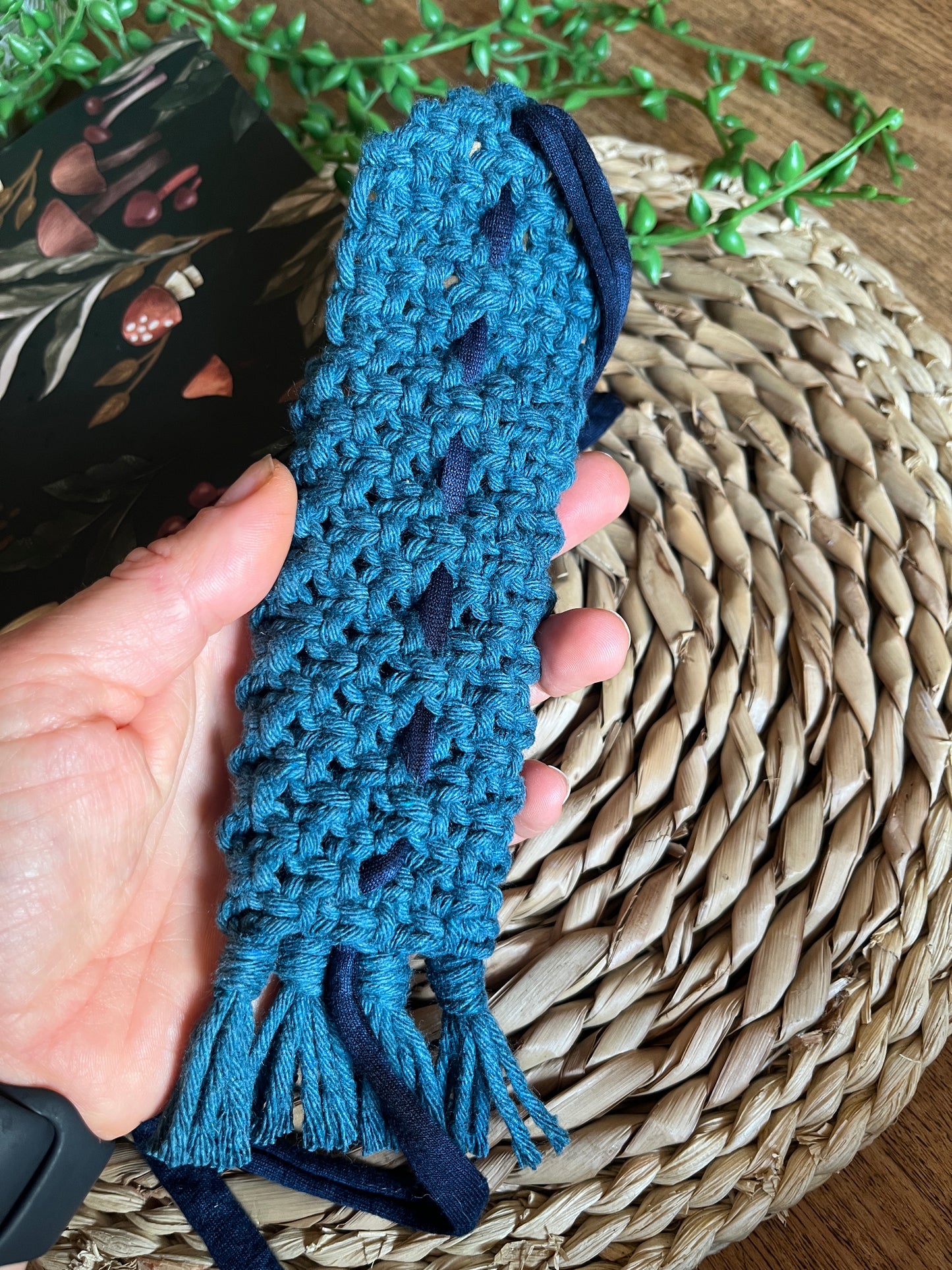 A hand holding a handmade macrame wrap around pen holder, made with petrol blue recycled cotton yarn. Displayed next a notebook which has a mushroom pattern, on a wicker mat. The photo shows the reverse side of the pen holder, showing the intricate yarn knots.