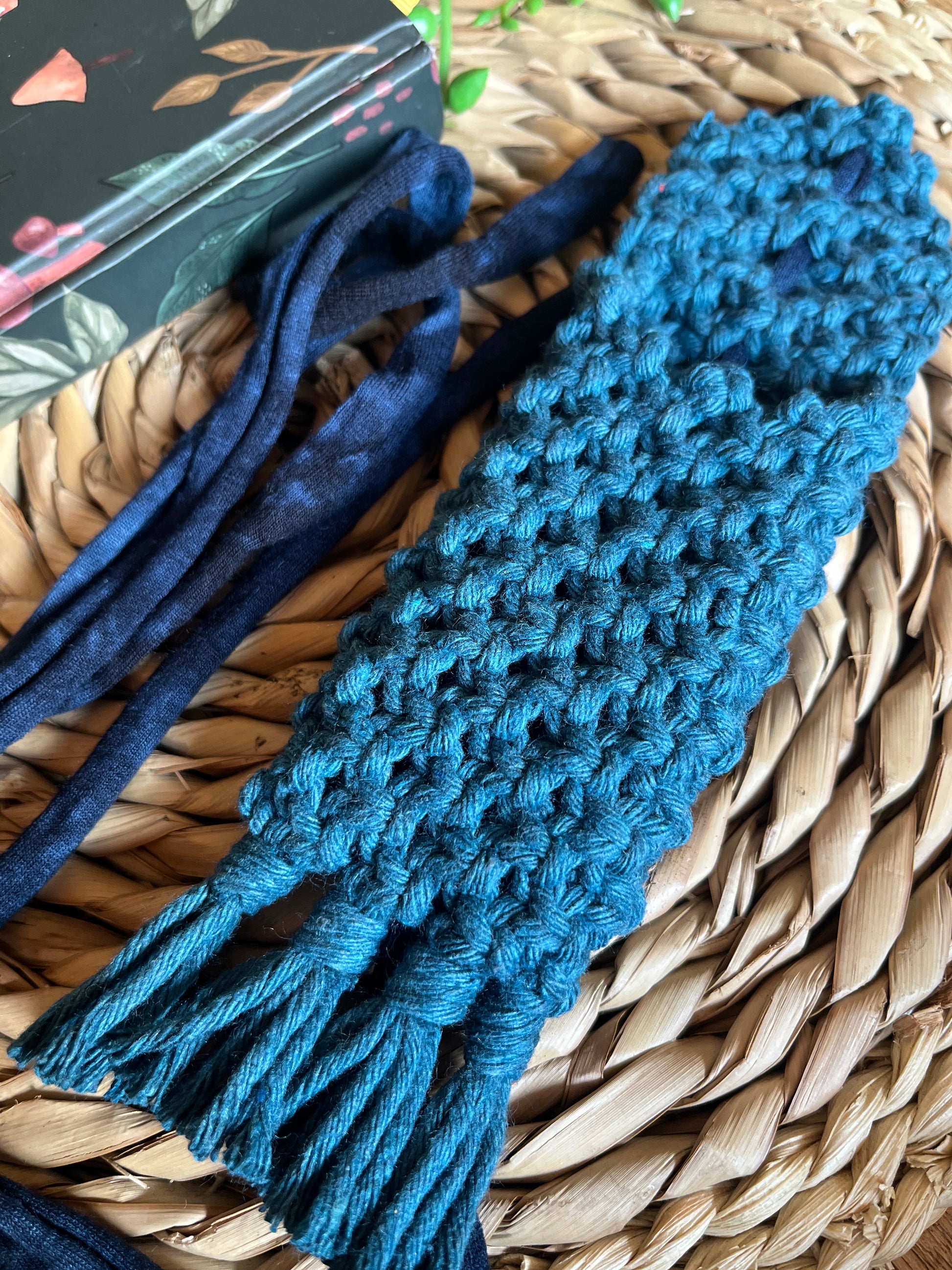 Handmade macrame wrap around pen holder, made with petrol blue recycled cotton yarn. Displayed next a notebook which has a mushroom pattern, on a wicker mat.