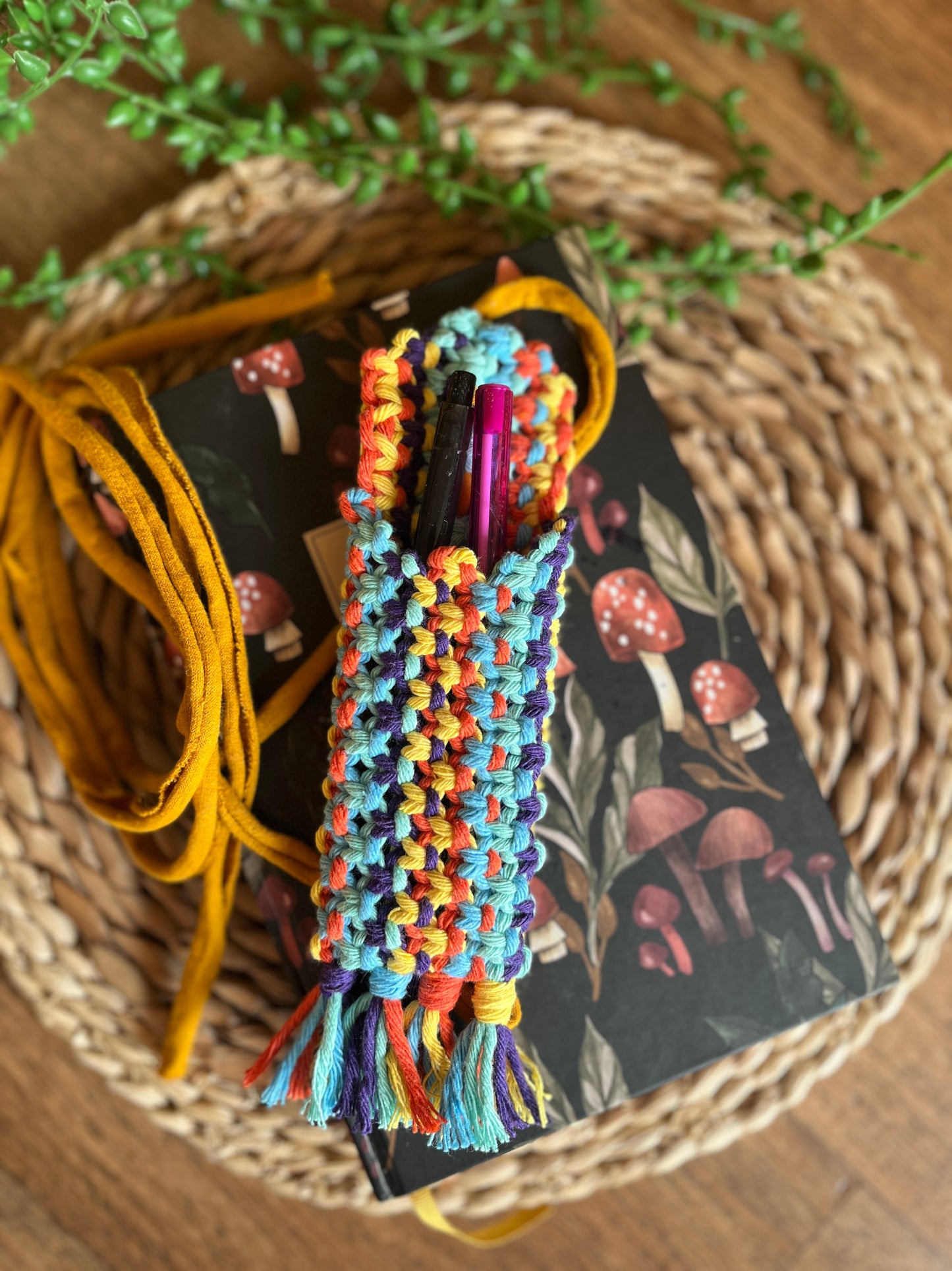 Handmade macrame wrap around pen holder, made with multiple colours of recycled cotton yarn. Displayed on a notebook which has a mushroom pattern, on a wicker mat. The pen holder has two pens inside of it, one black and one pink.