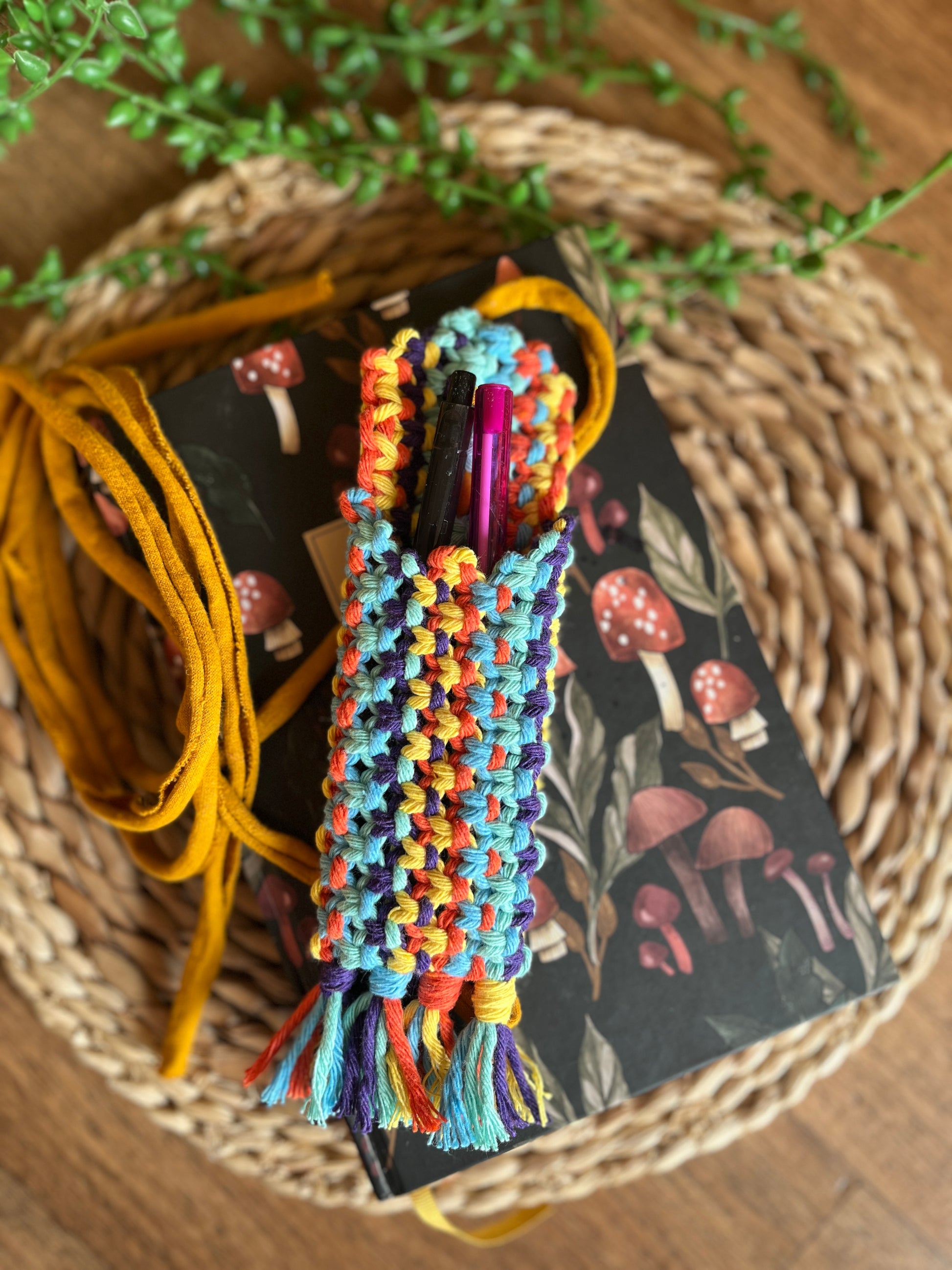 Handmade macrame wrap around pen holder, made with multiple colours of recycled cotton yarn. Displayed on a notebook which has a mushroom pattern, on a wicker mat. The pen holder has two pens inside of it, one black and one pink.