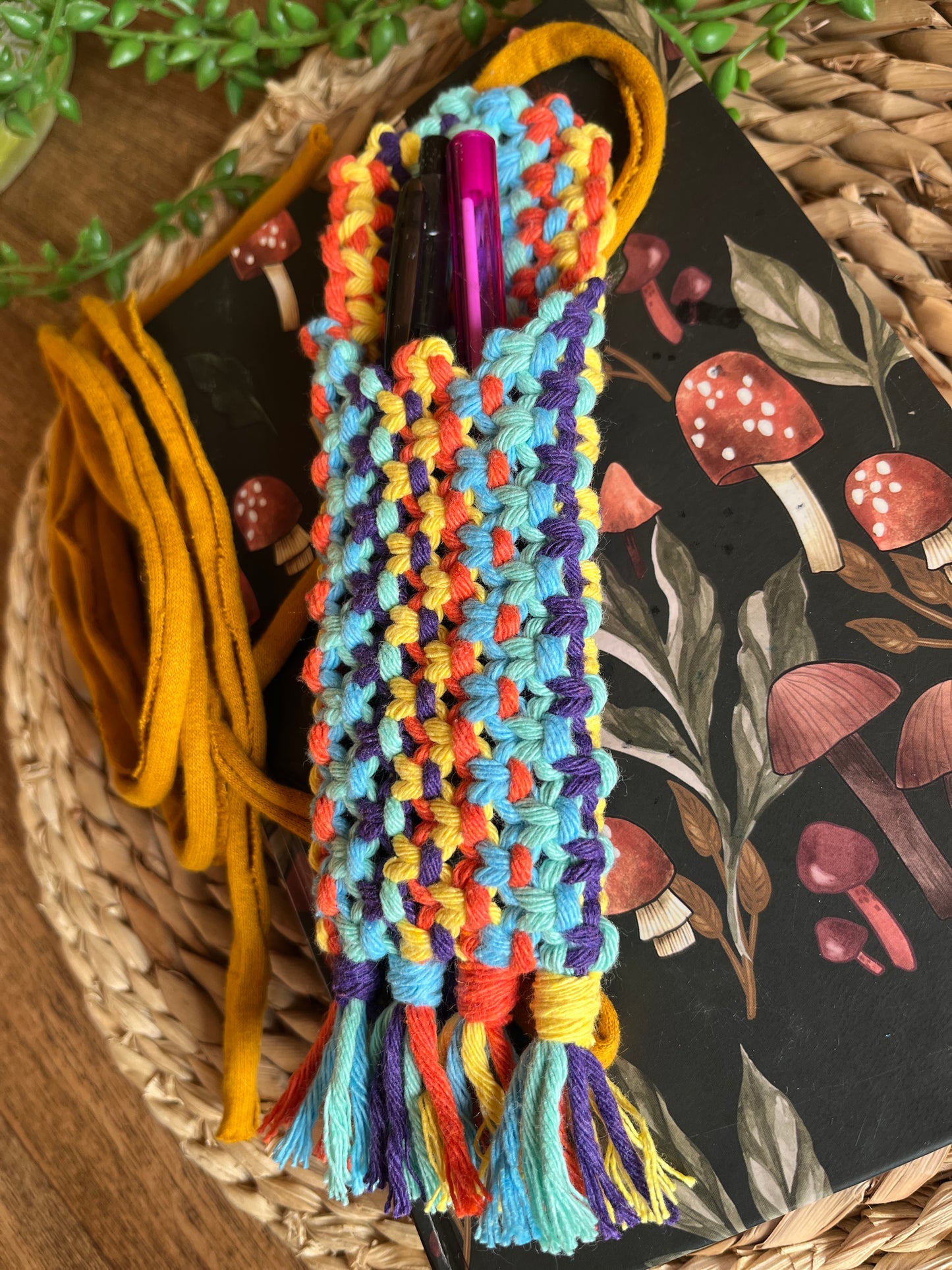 Handmade macrame wrap around pen holder, made with multiple colours of recycled cotton yarn. Displayed on a notebook which has a mushroom pattern, on a wicker mat. The pen holder has two pens inside of it, one black and one pink.