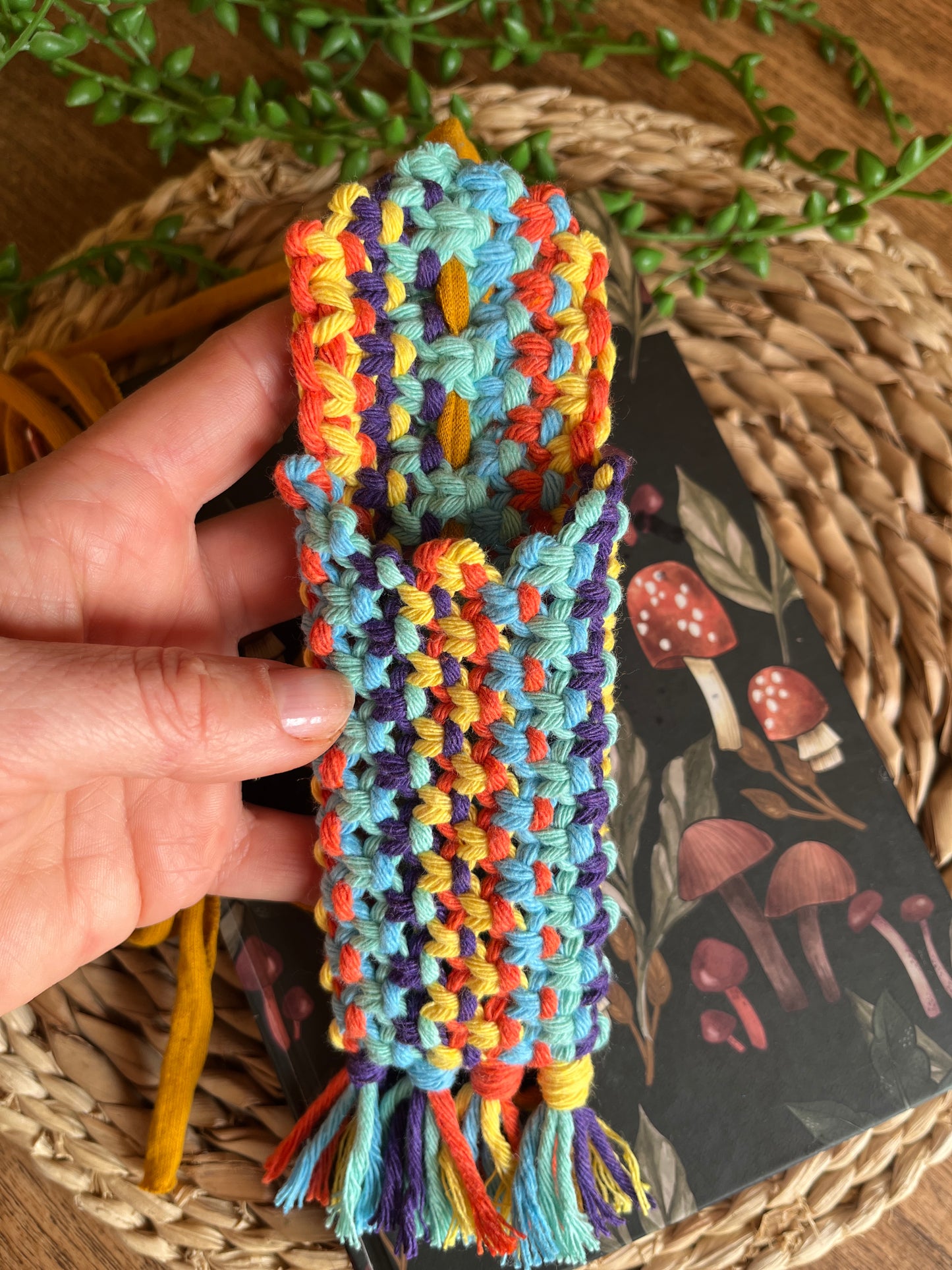 A hand holding a handmade macrame wrap around pen holder, made with multiple colours of recycled cotton yarn. Displayed on a notebook which has a mushroom pattern, on a wicker mat. The pen holder has two pens inside of it, one black and one pink.