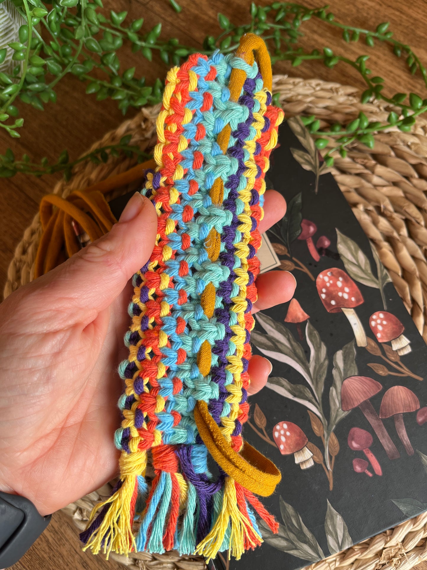 A hand holding a handmade macrame wrap around pen holder, made with multiple colours of recycled cotton yarn. Displayed on a notebook which has a mushroom pattern, on a wicker mat. The image shows the intricate knots on the back of the pen holder.
