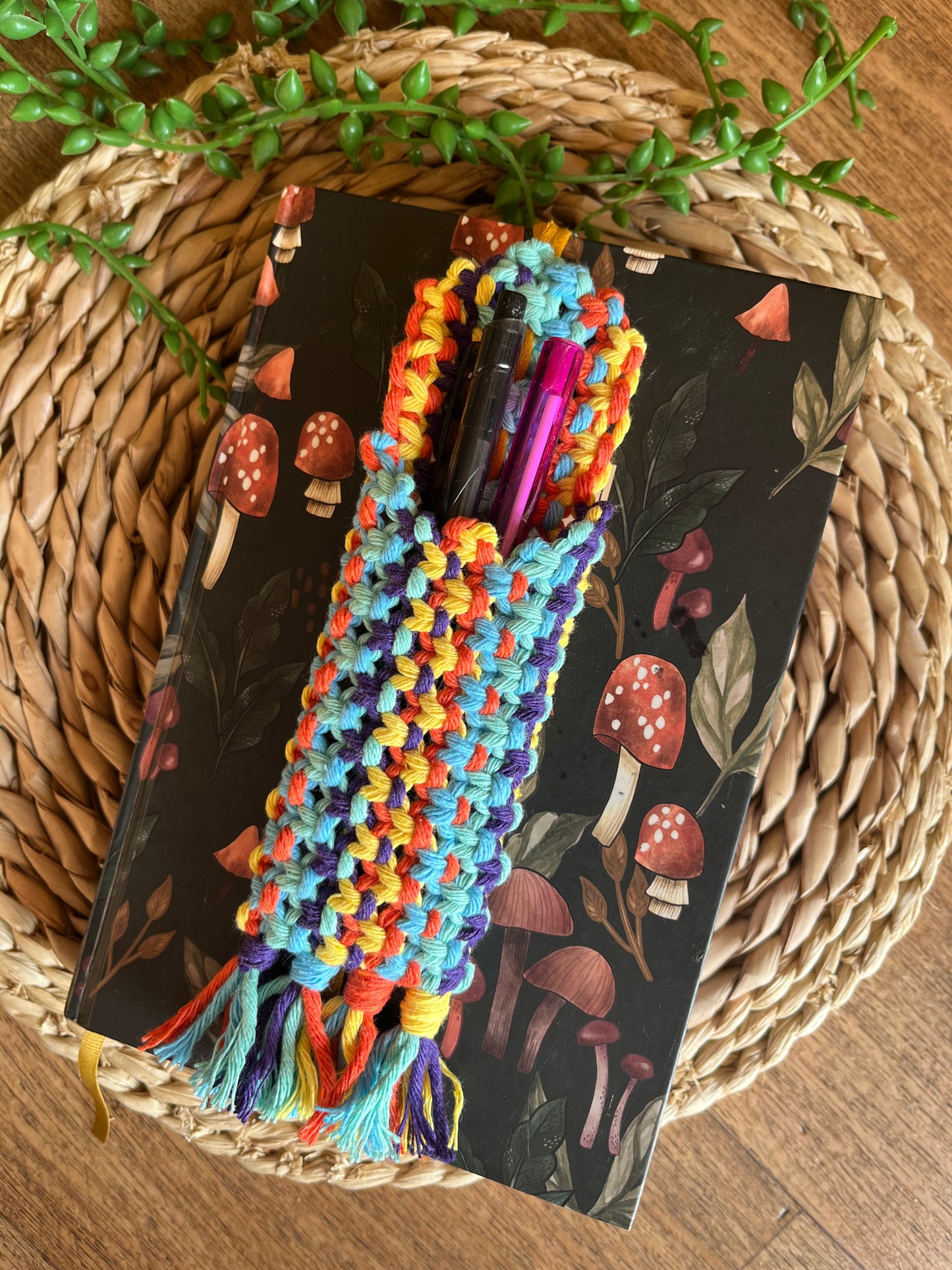 Handmade macrame wrap around pen holder, made with multiple colours of recycled cotton yarn. Displayed on a notebook which has a mushroom pattern, on a wicker mat. The pen holder has two pens inside of it, one black and one pink.