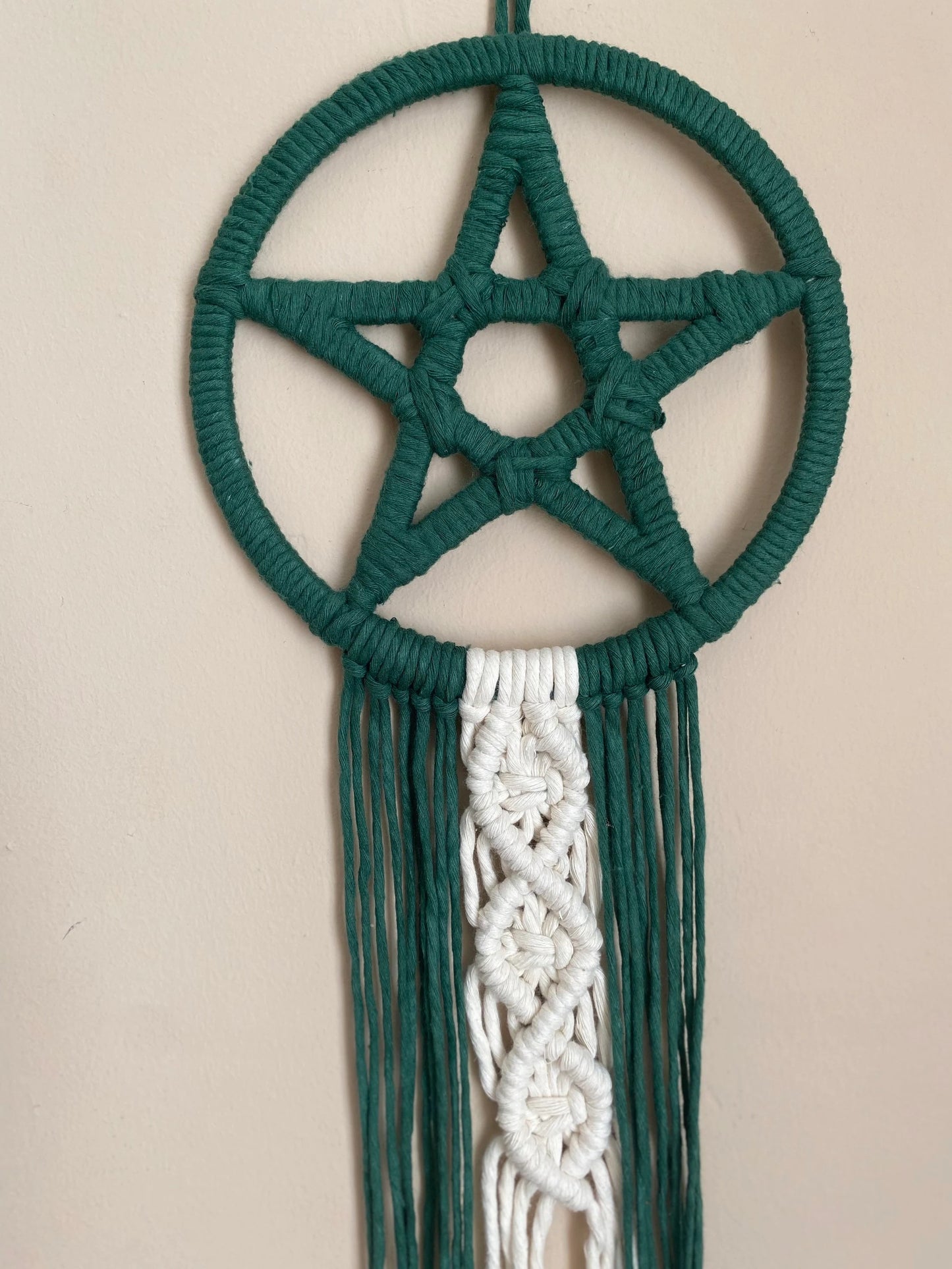 Handmade macrame pentagram wall hanging made with green and natural flowing tails, hanging against a beige wall.