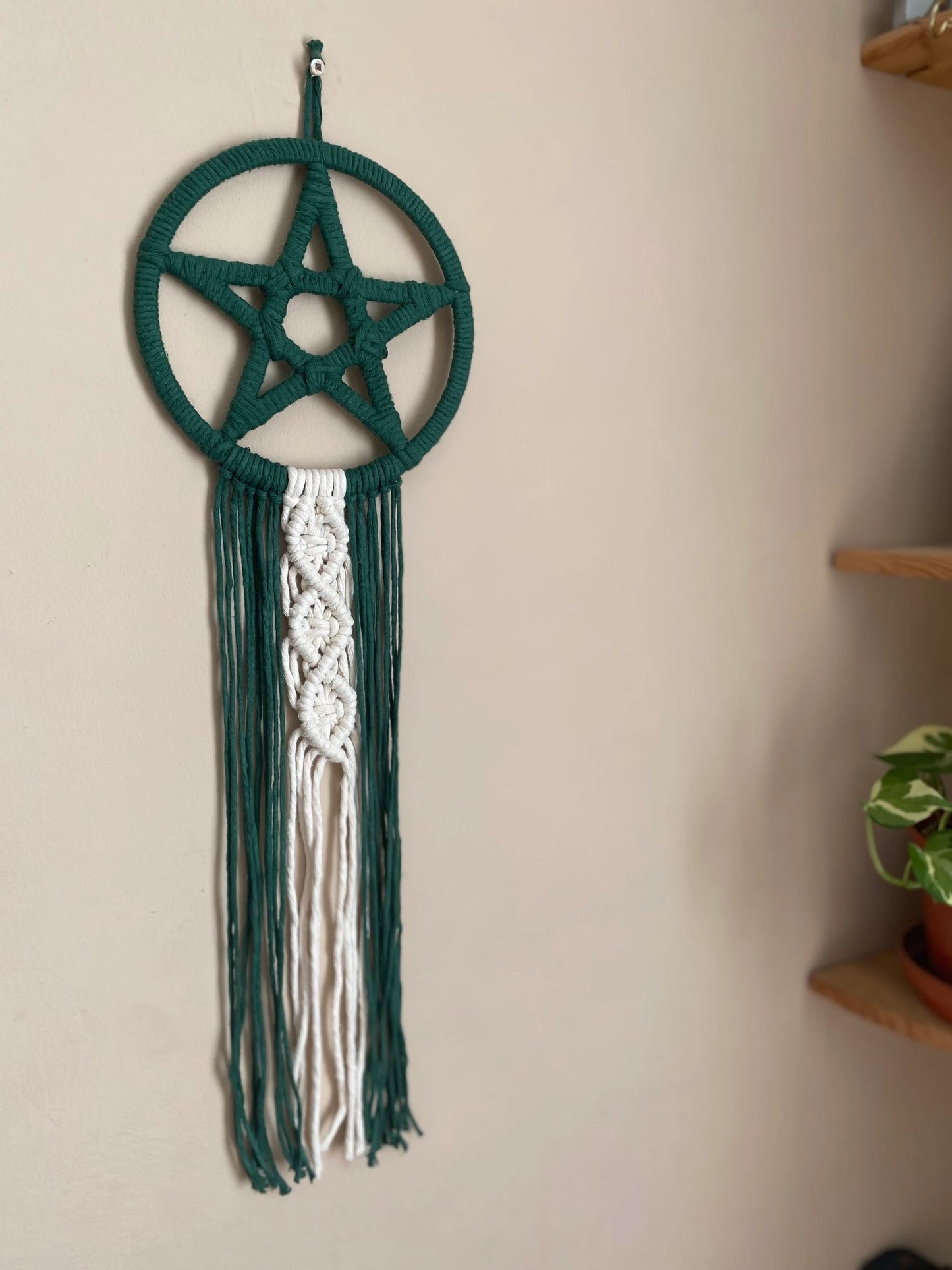Handmade macrame pentagram wall hanging made with green and natural yarns with long flowing tails, hanging against a beige wall next to a set of corner shelves.
