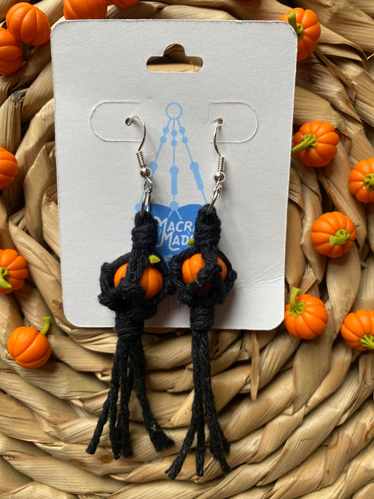 A pair of earrings made with small orange pumpkins wrapped in black yarn on silver hooks, displayed on a white backing card with a wicker mat and lots of small orange pumpkins in the background.