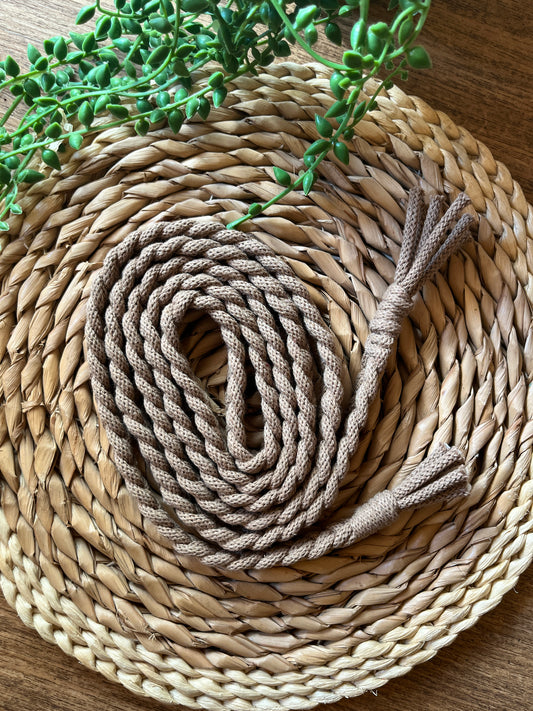 A coiled beige, braided rope belt with tassels at both ends, placed on a round wicker surface.