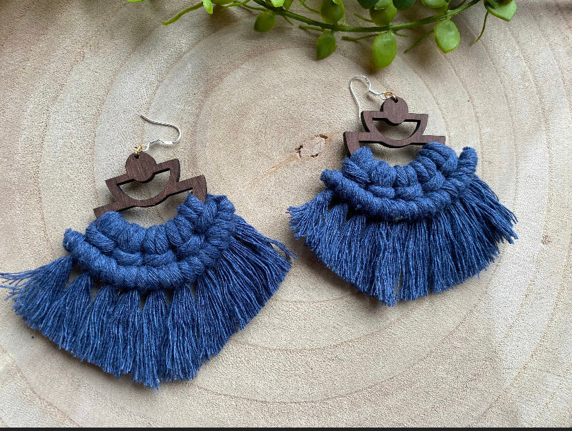 A pair of handmade macrame earrings made with blue yarn, on wooden semi circle findings with silver hooks, and neat fringed detail, placed on a wooden surface surrounded by greenery.