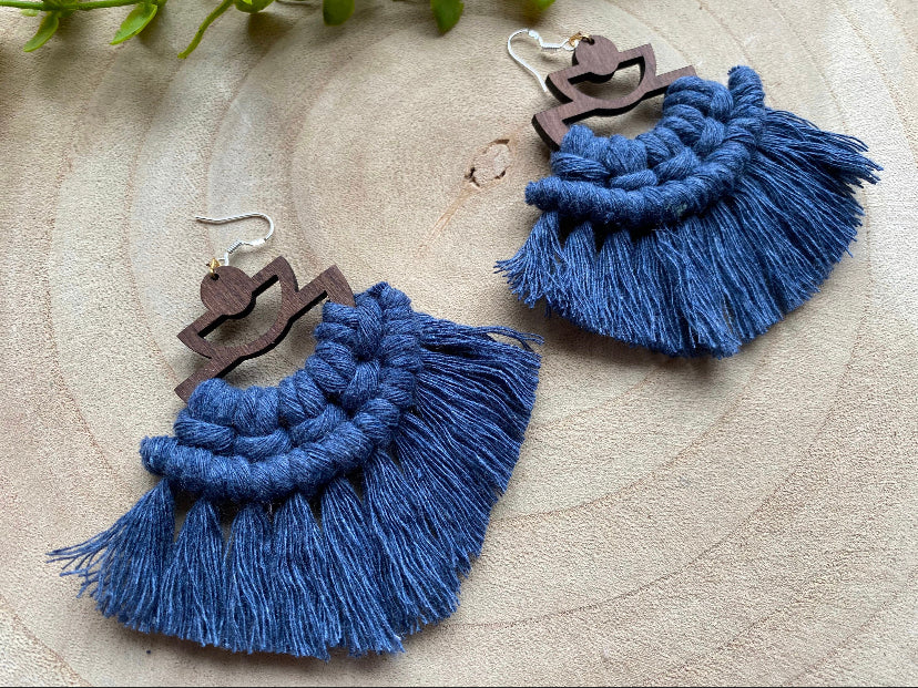 A pair of handmade macrame earrings made with blue yarn, on wooden semi circle findings with silver hooks, and neat fringed detail, placed on a wooden surface.