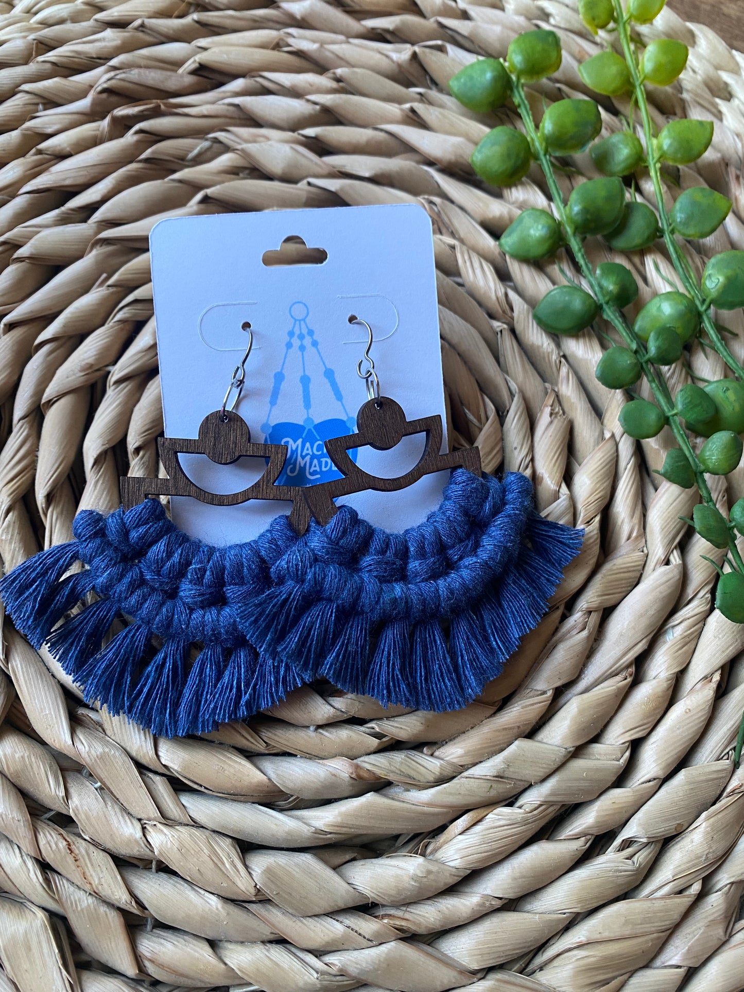 A pair of handmade macrame earrings made with blue yarn, on wooden semi circle findings with silver hooks, and neat fringed detail, on a white backing card.