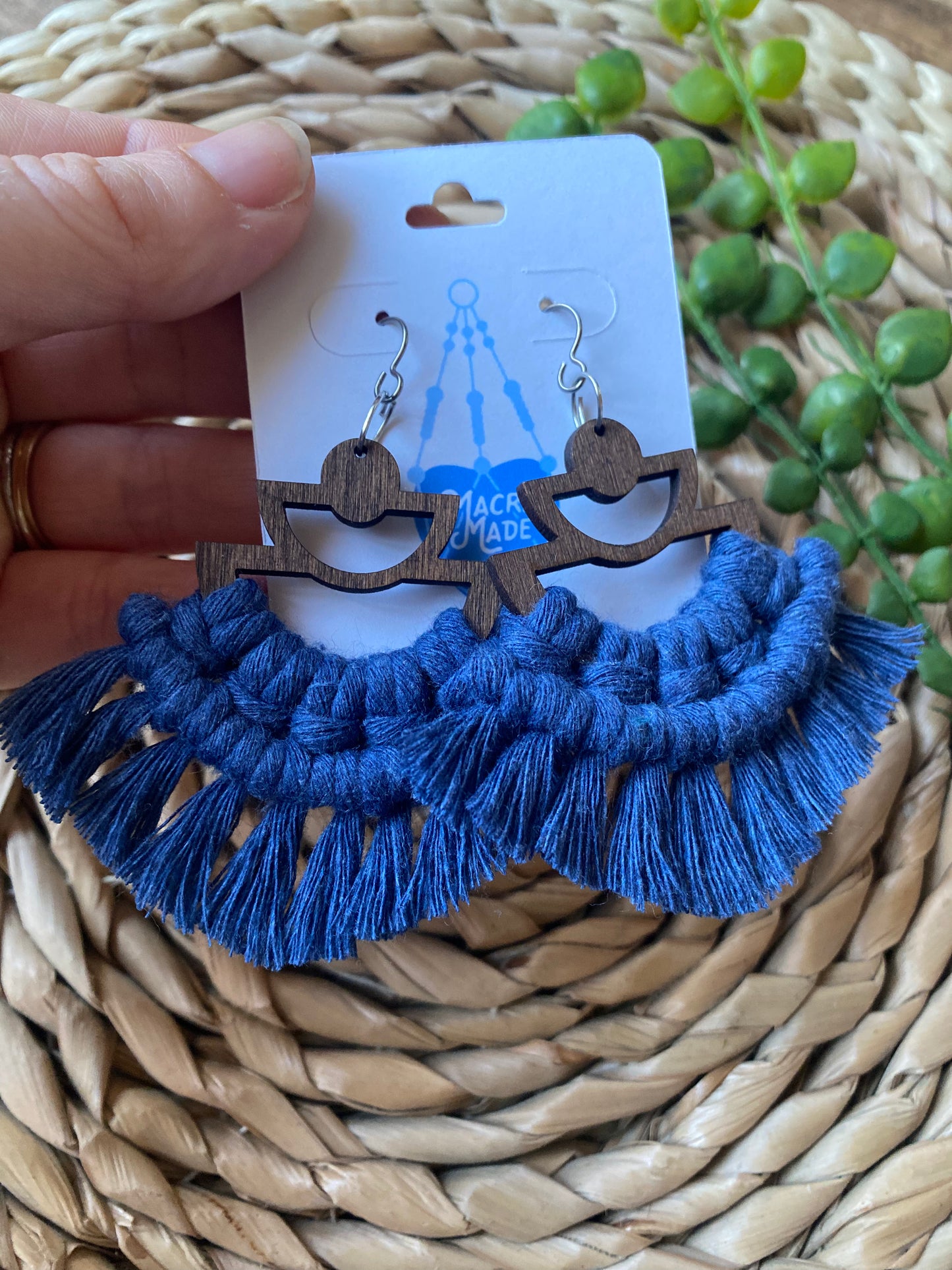 A hand holding a pair of handmade macrame earrings made with blue yarn, on wooden semi circle findings with silver hooks, and neat fringed detail, attached to a white backing card.
