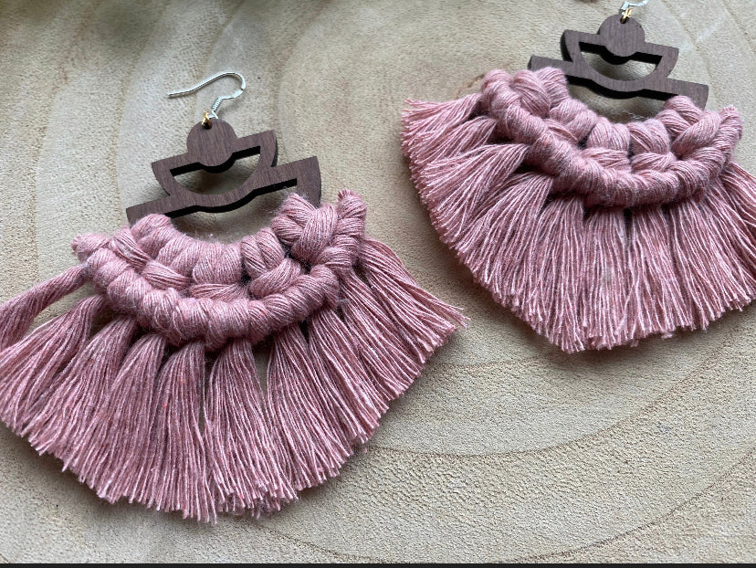 A pair of handmade macrame earrings made with blush pink yarn, on wooden semi circle findings with silver hooks, and neat fringed detail, placed on a wooden surface.