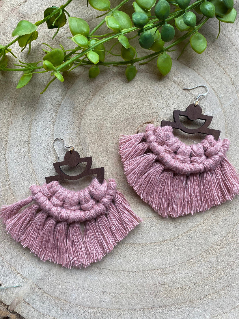 A pair of handmade macrame earrings made with blush pink yarn, on wooden semi circle findings with silver hooks, and neat fringed detail, placed on a wooden surface surrounded by greenery.