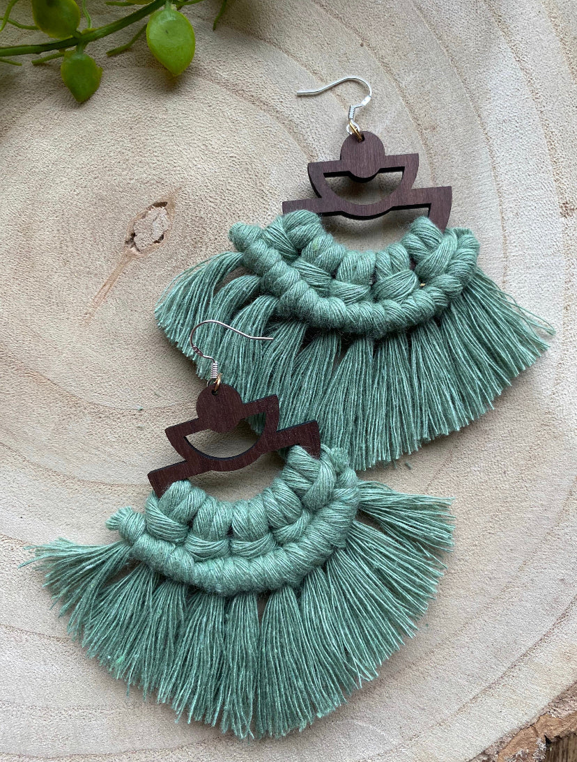 A pair of handmade macrame earrings made with green yarn, on wooden semi circle findings with silver hooks, and neat fringed detail, placed on a wooden surface.