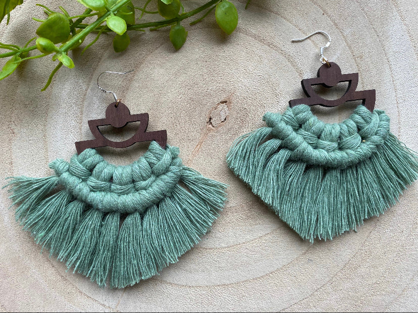A pair of handmade macrame earrings made with green yarn, on wooden semi circle findings with silver hooks, and neat fringed detail, placed on a wooden surface surrounded by greenery.