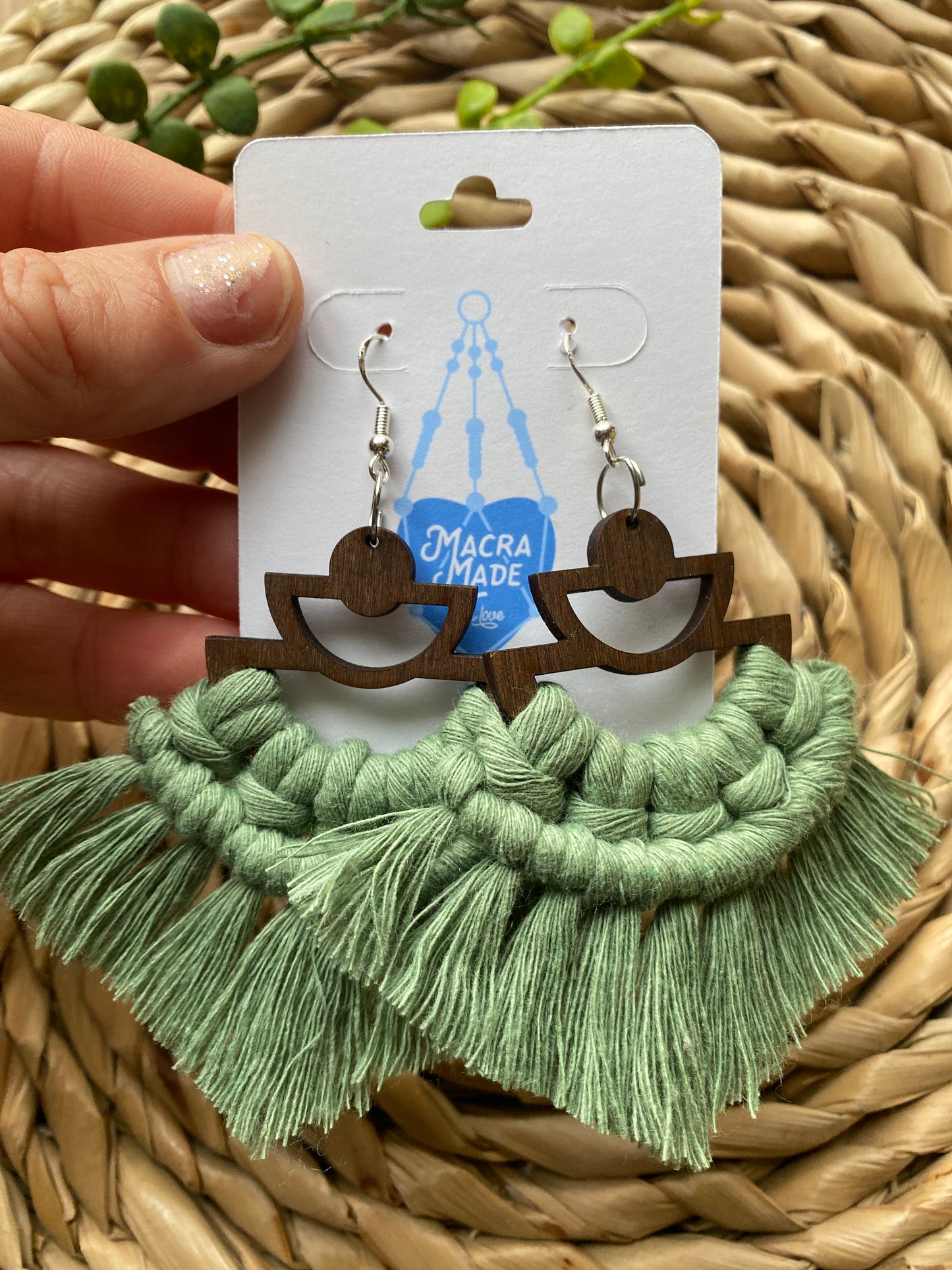 A hand holding a pair of handmade macrame earrings made with green yarn, on wooden semi circle findings with silver hooks, attached to a white backing card.