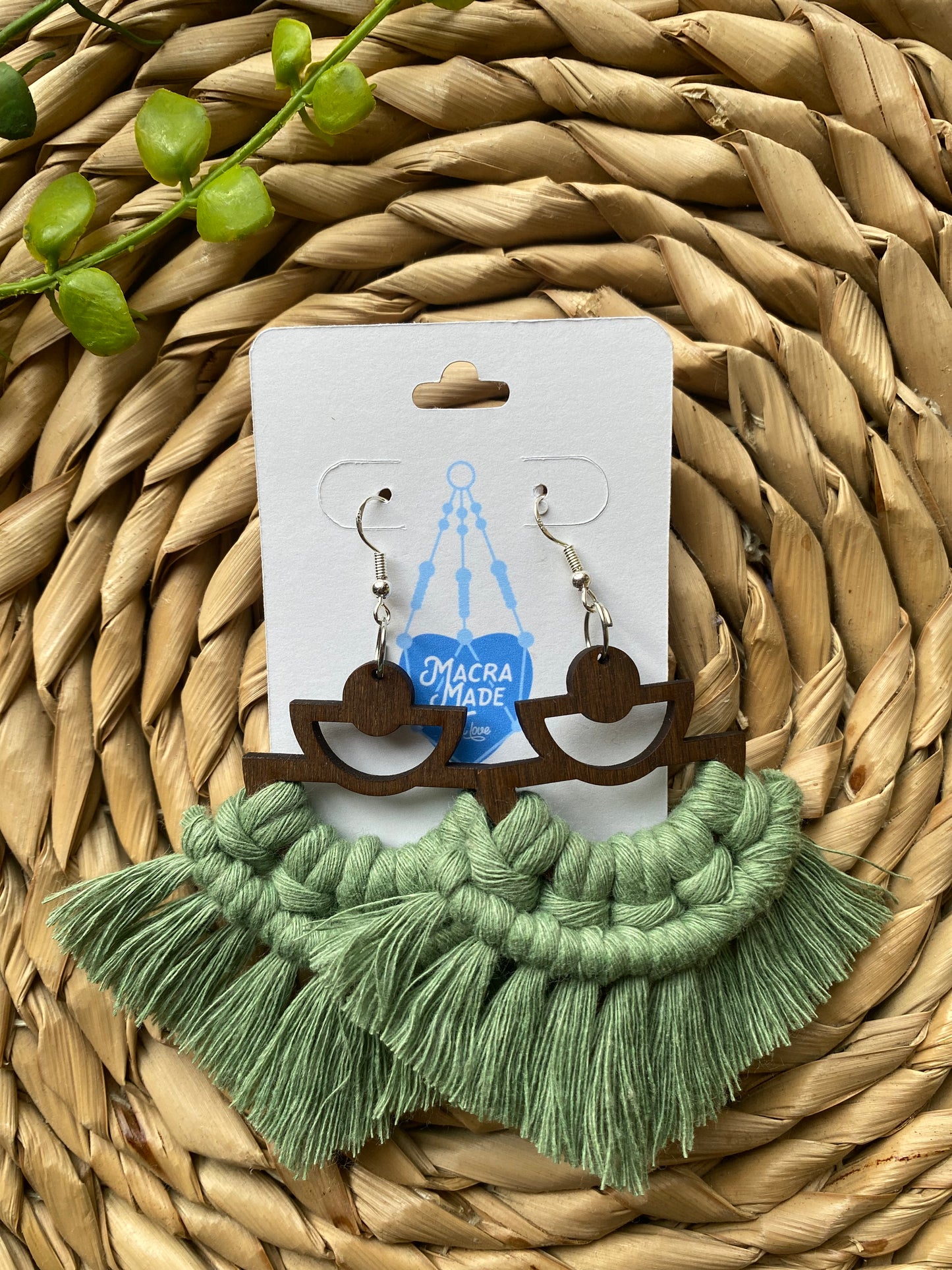 A pair of handmade macrame earrings made with green yarn, on wooden semi circle findings with silver hooks, attached to a white backing card.