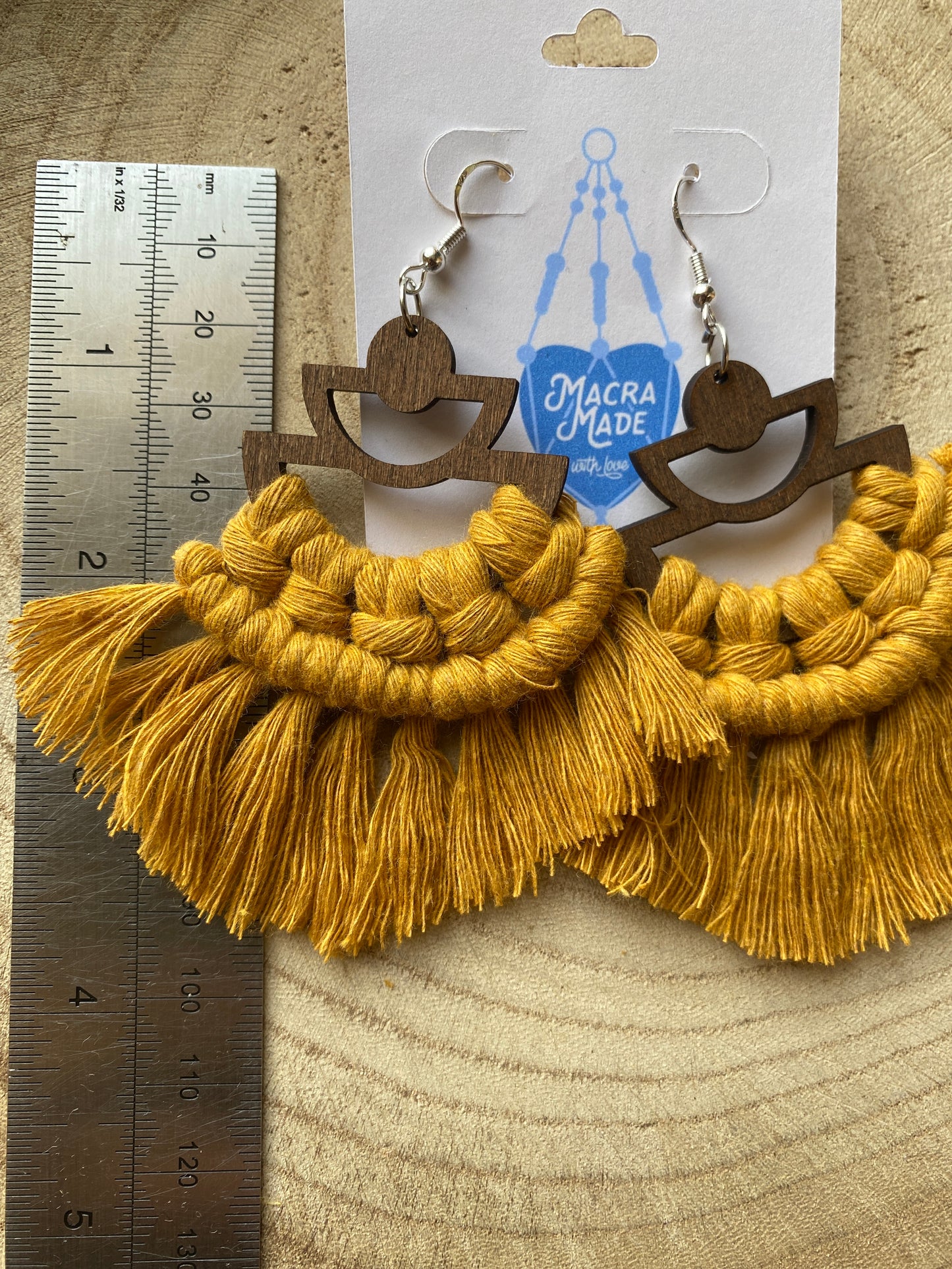 A pair of handmade macrame earrings made with mustard yellow yarn, on wooden semi circle findings with silver hooks, and neat fringed detail, placed on a wooden surface next to a metal rule