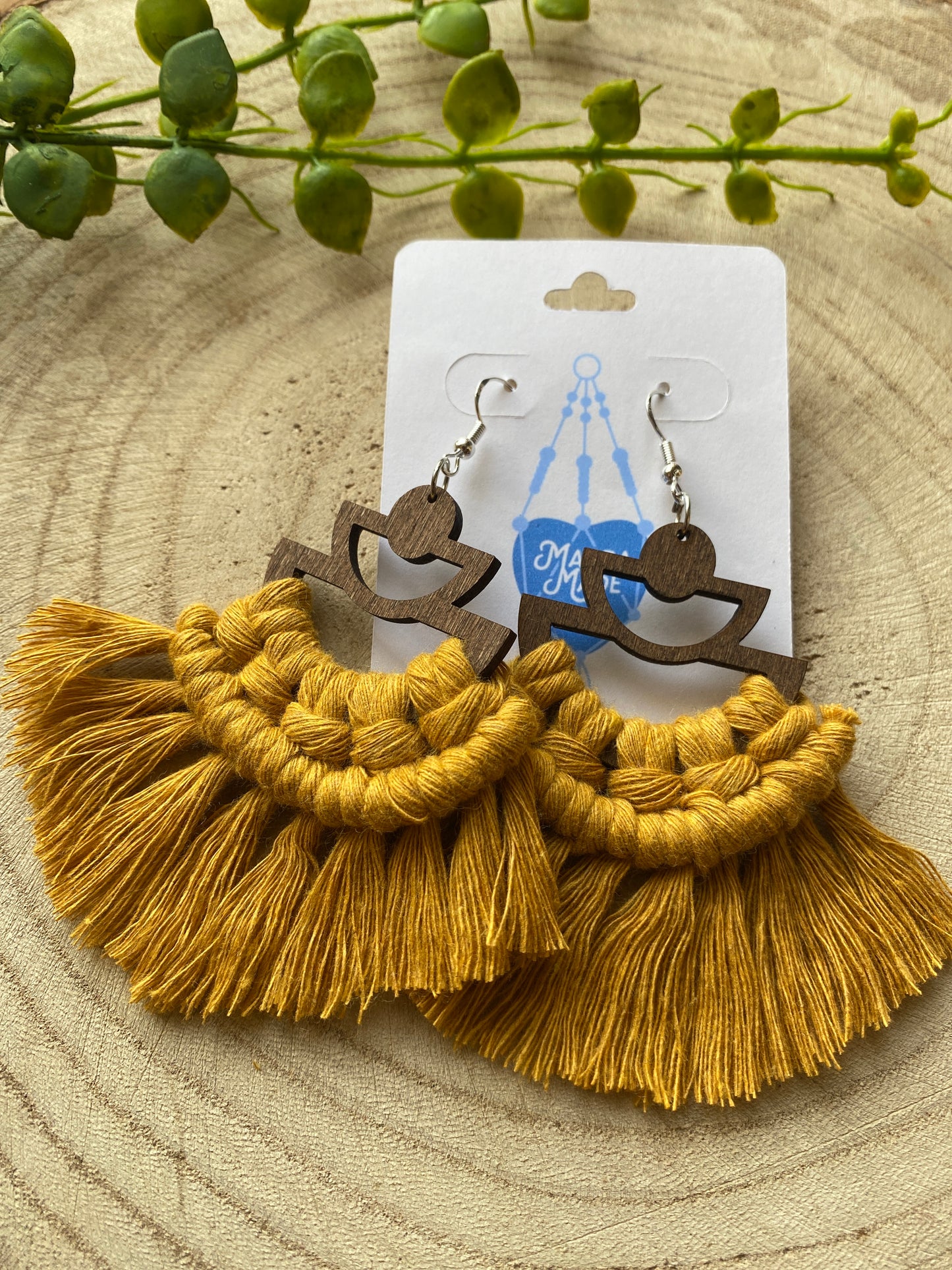 A pair of handmade macrame earrings made with mustard yellow yarn, on wooden semi circle findings with silver hooks, attached to a white backing card, and placed on a wooden board.