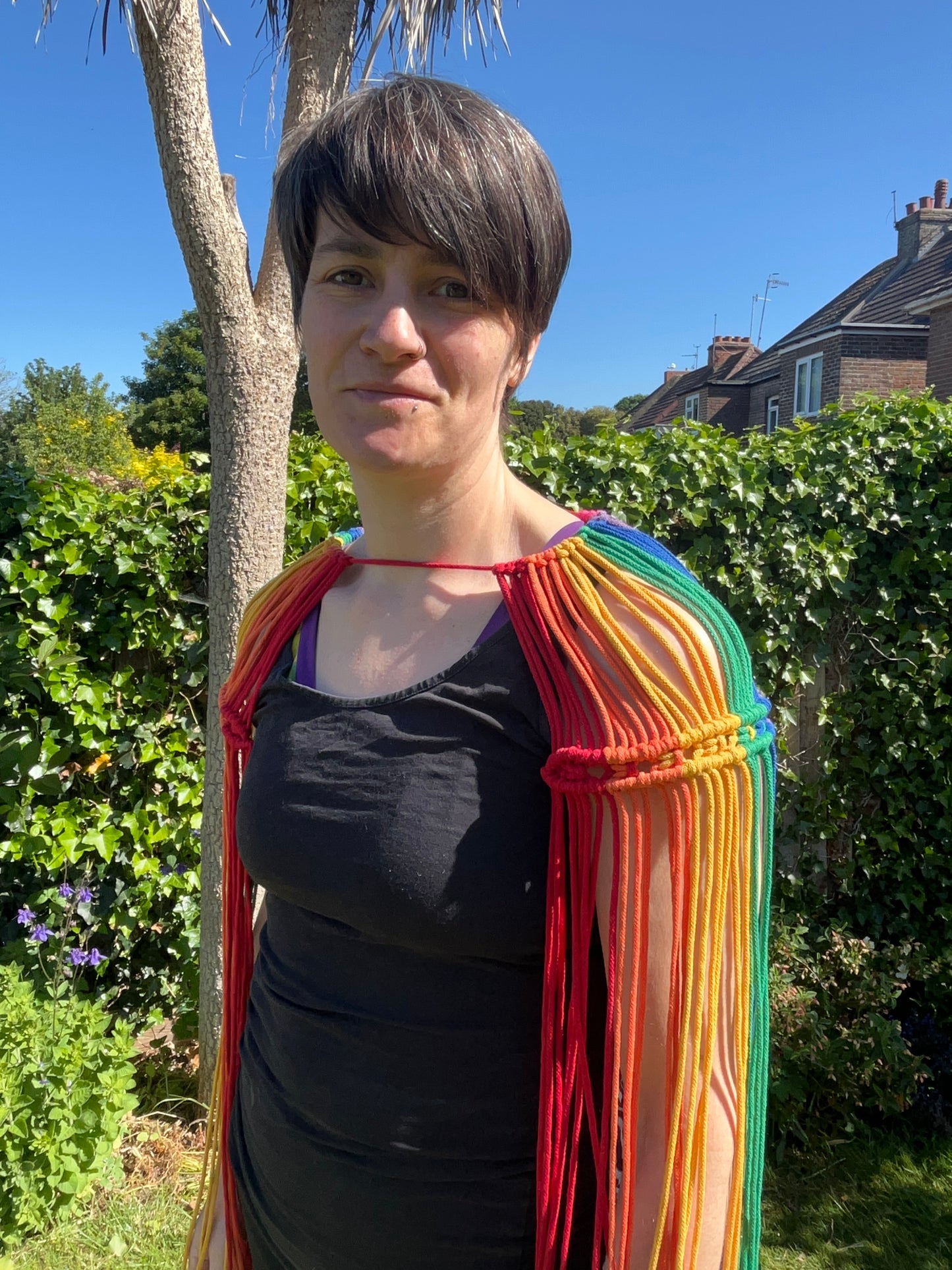 A female wearing a black vest, and handmade macrame shoulder accessory epaulette, stands outside on a sunny day, smiling towards the camera.