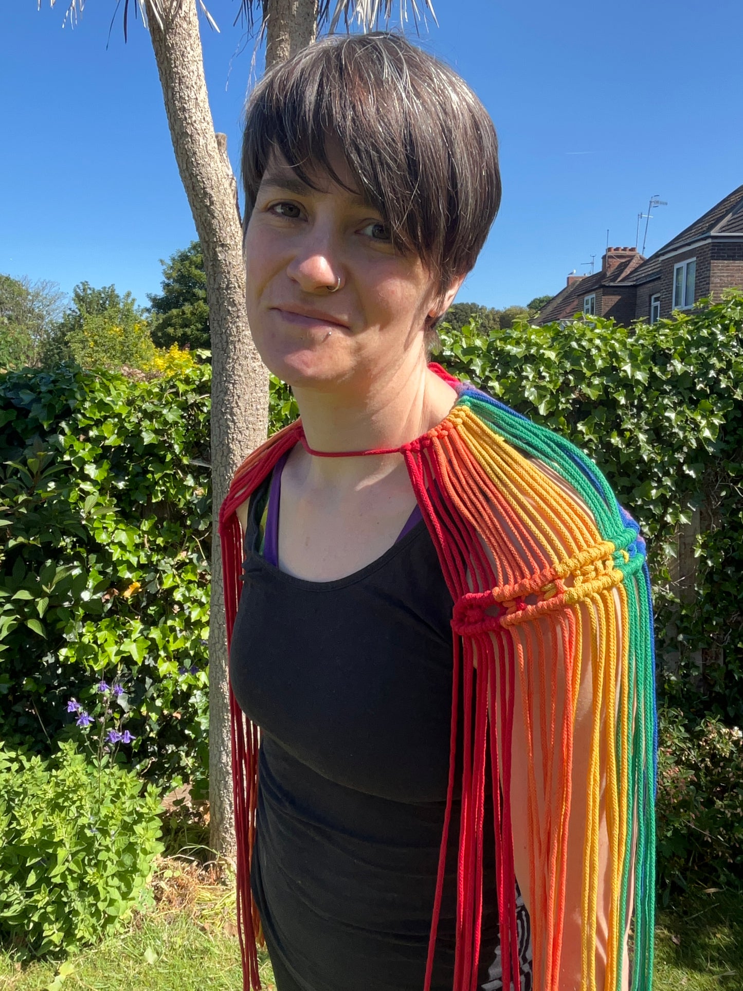 A female wearing a black vest, and handmade macrame shoulder accessory epaulette, stands outside on a sunny day, smiling towards the camera.