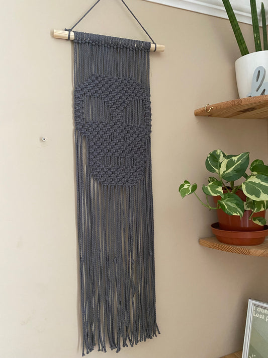 Long dark grey macrame wall hanging with a knotted pattern in the shape of a skull, made on a wooden dowel, hanging against a beige wall next to a small houseplant on a corner shelf.