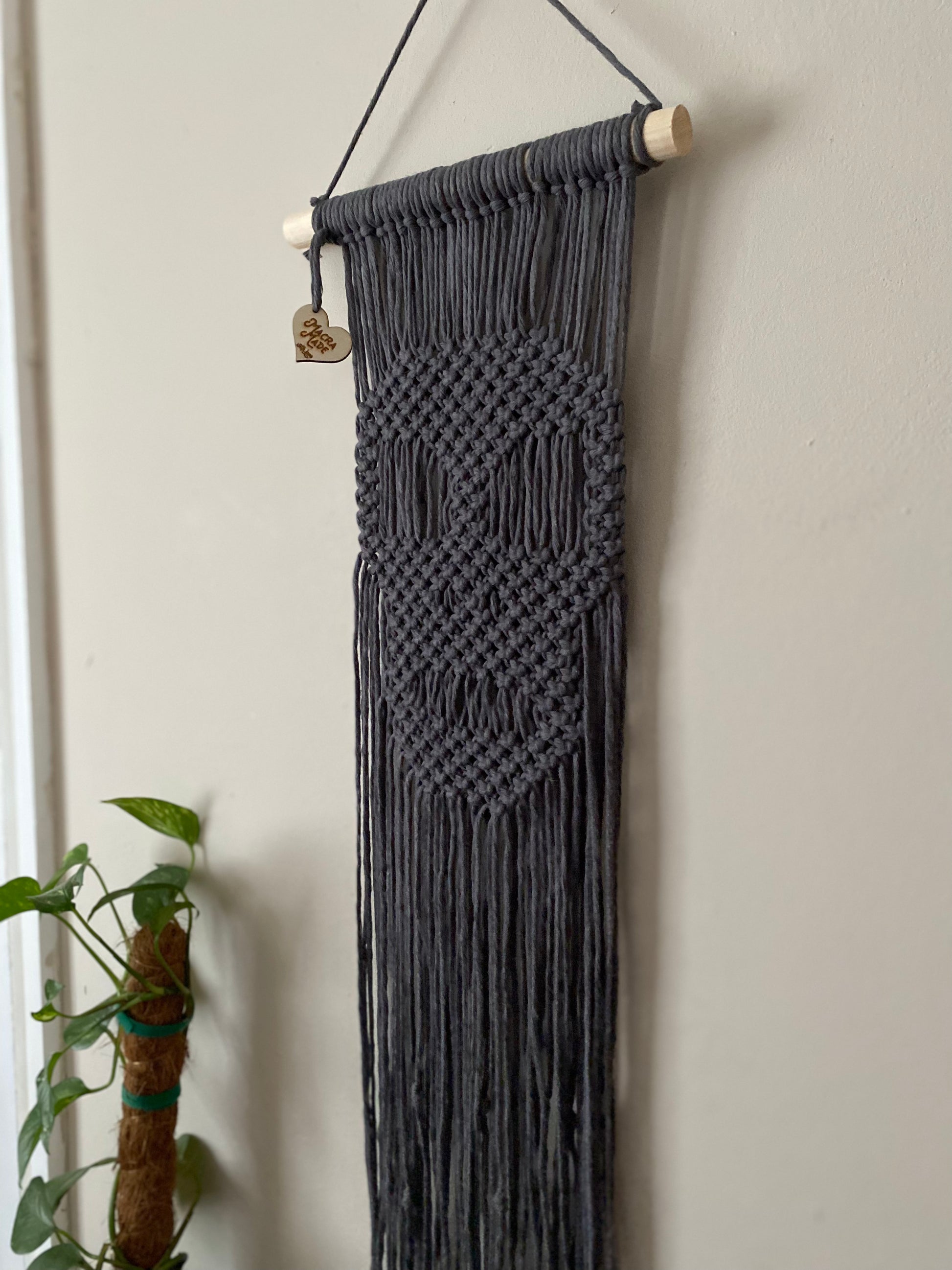 Close up of a long dark grey macrame wall hanging with a knotted pattern in the shape of a skull, made on a wooden dowel, hanging against a beige wall.