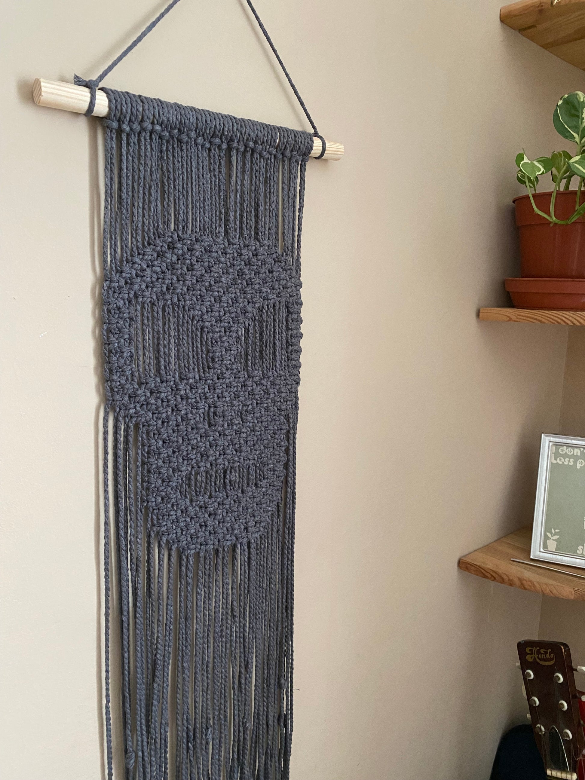 Dark grey macrame wall hanging with a knotted pattern in the shape of a skull, made on a wooden dowel, hanging against a beige wall next to a small houseplant on a corner shelf.